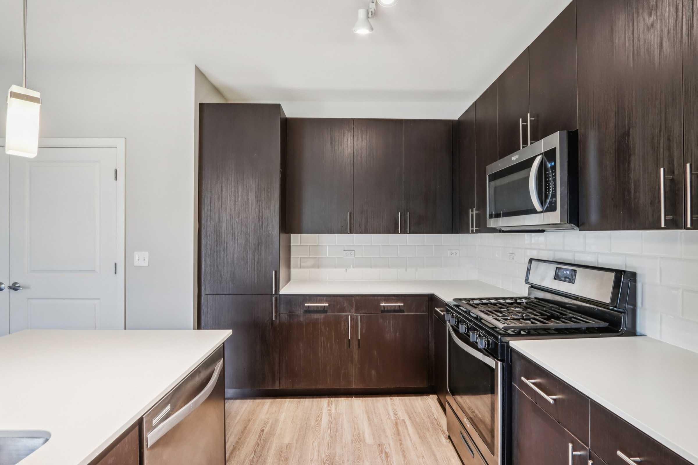 Modern kitchen featuring dark wood cabinetry, stainless steel appliances including a microwave and stove, and a white countertop. The space has a light color scheme with a glass pendant light and a door leading to another room.