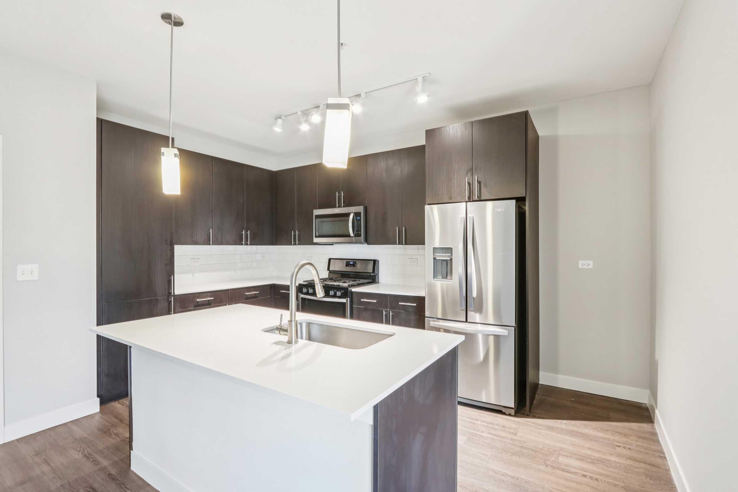 Modern kitchen featuring dark wooden cabinets, stainless steel appliances, a large island with a white countertop, and pendant lighting. The space is open and bright, with a seamless flow between cabinetry and the stainless steel fridge and stove.