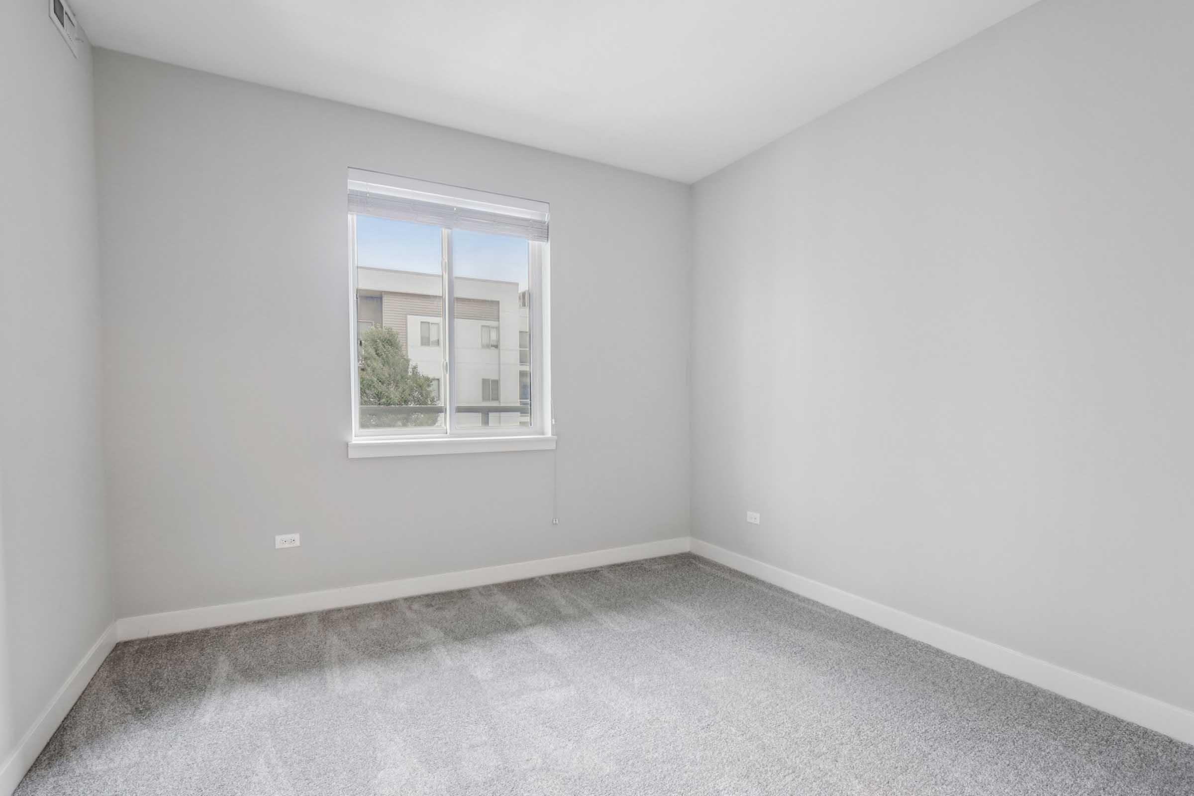 An empty room with light gray walls and carpeted flooring. A window with a white frame is located on the left side, allowing natural light to enter. The overall space appears clean, bright, and modern, suitable for various purposes or personal decoration.