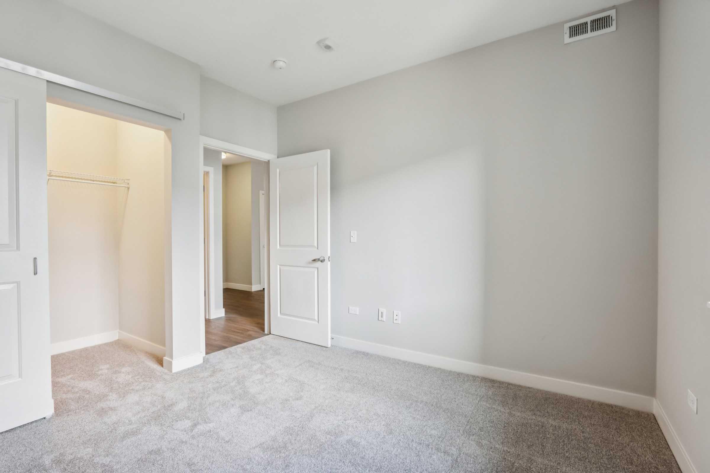 A partially furnished room with light gray walls and carpeted flooring. It features a single door leading to a closet space and another door opening to an adjacent area. The room is well-lit with natural light coming in.
