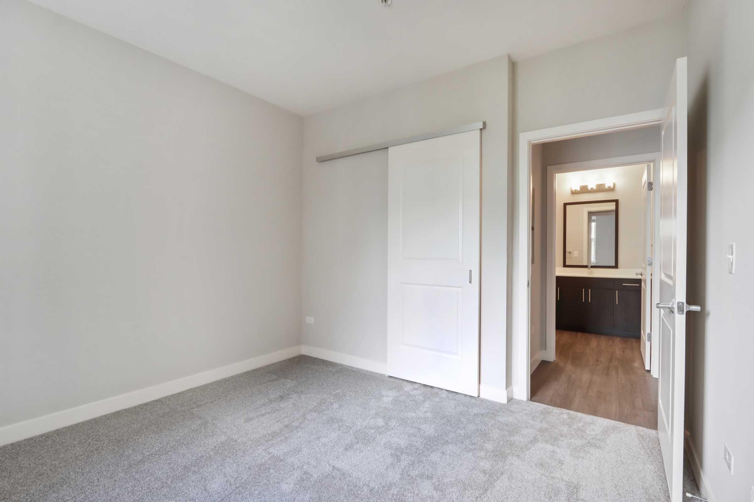 Interior of a room featuring light gray walls and plush carpet. A large sliding door is positioned on the left, leading to an adjoining bathroom that is partially visible. The bathroom has modern fixtures and a mirror above the vanity, with cabinetry in dark tones.