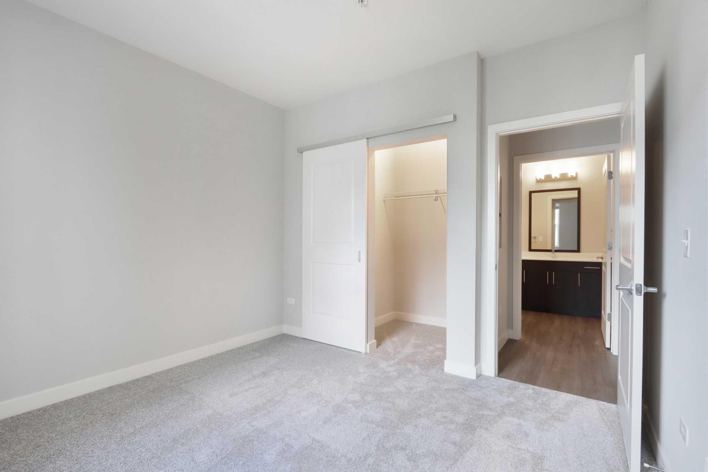 A spacious, empty bedroom with light gray walls and carpet. There is a sliding door leading to a closet and an adjoining doorway that opens to a bathroom area with a modern vanity and mirror. The overall atmosphere is bright and inviting, ideal for personal decoration.
