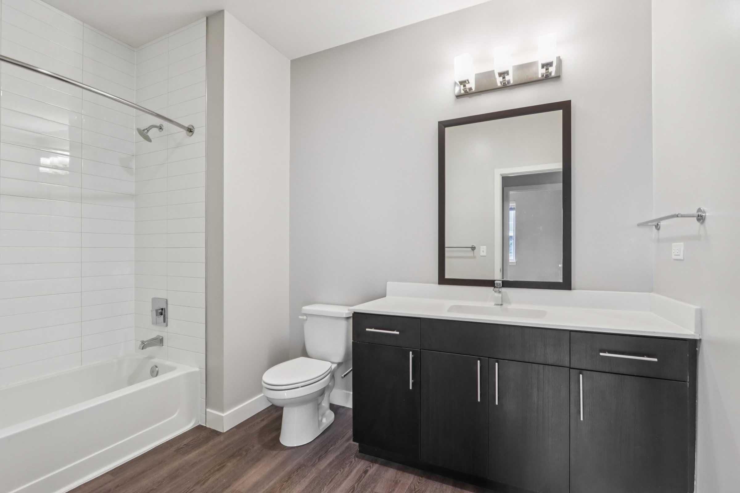 Modern bathroom featuring a bathtub with a showerhead, a toilet, and a sleek dual-sink vanity with dark cabinets. A large mirror hangs above the vanity, and the walls are painted in a neutral color. The floor is adorned with wood-like tiles, creating a warm and inviting atmosphere.