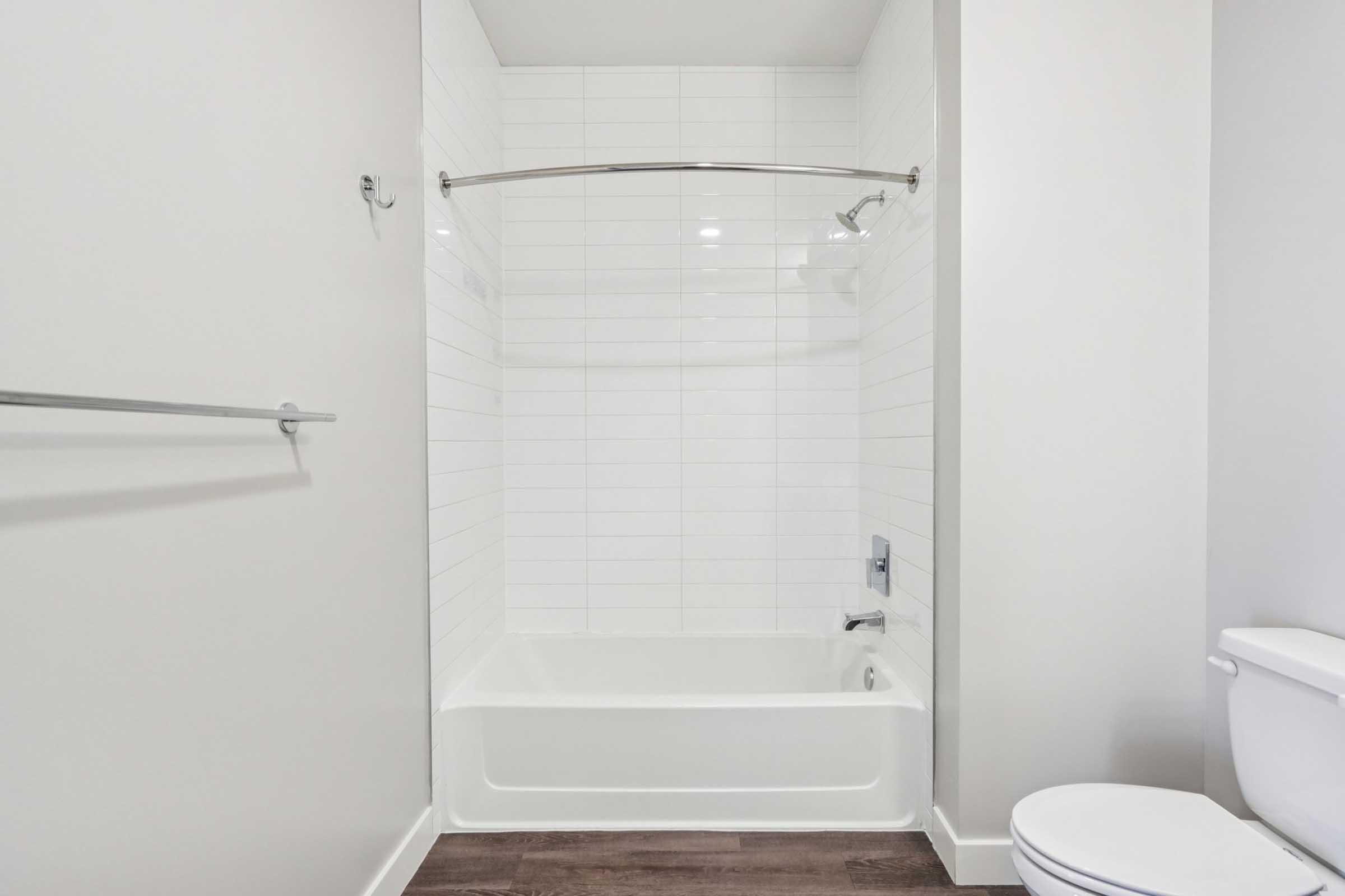 A clean, modern bathroom featuring a white tiled shower/bathtub combination with a chrome shower rod, a simple white toilet, and dark wooden flooring. The walls are painted a light color, enhancing the spacious feel of the room.