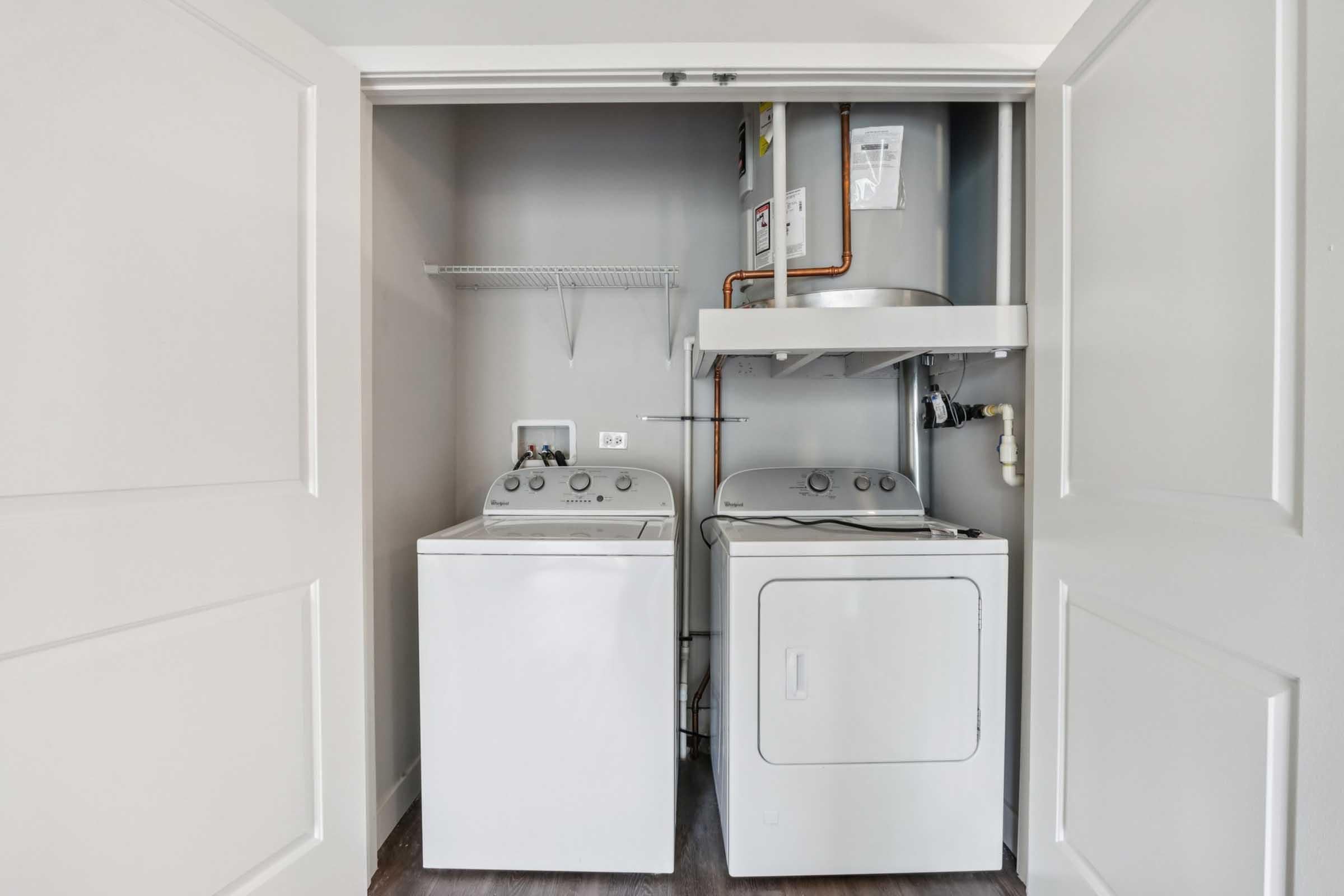 A small laundry room featuring a washer and dryer side by side. The appliances are situated in a closet-like space with white walls and doors. Above the washer, there is a shelf for storage, and a water heater is visible on the side, along with plumbing connections. The floor is a hardwood design.
