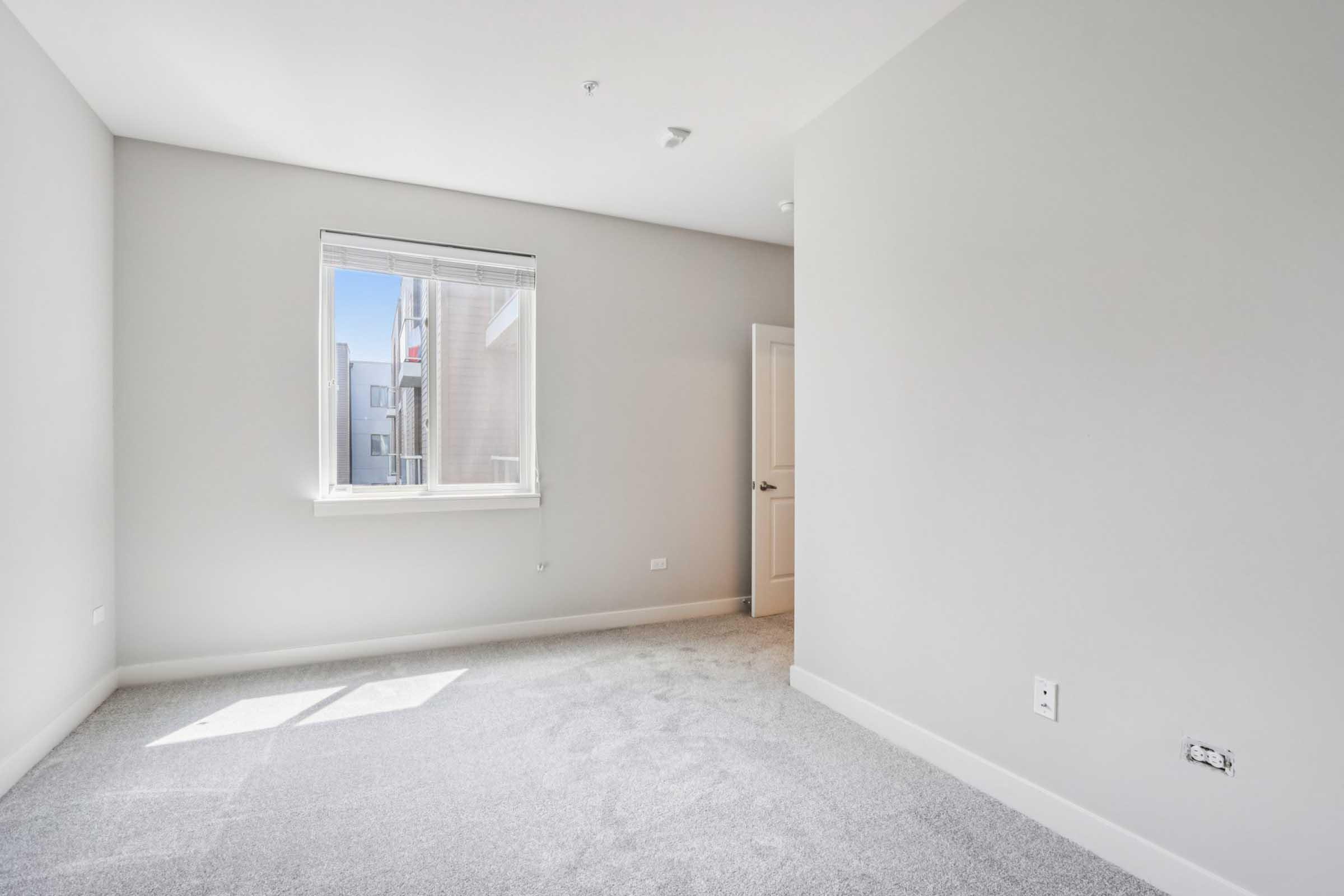 A bright, empty room with light gray walls and carpet. A window allows natural light to enter, illuminating the space. There is a closed door on the right and a small wall outlet visible. The overall atmosphere is clean and minimalistic, ideal for personalization.