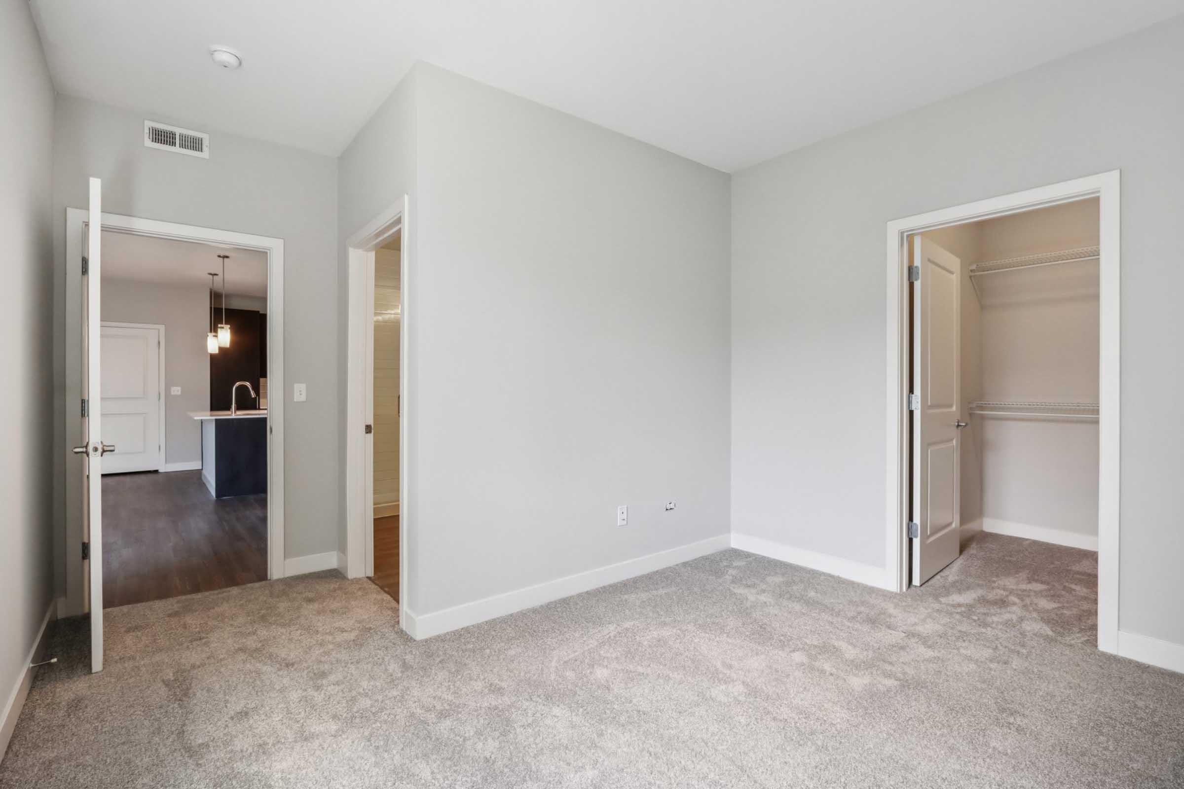 A spacious room with light gray walls and carpet flooring. There are two doorways: one leading to a closet and another opening to an adjacent room with a dark kitchen area visible in the background. Natural light illuminates the space, giving it a bright and airy feel.