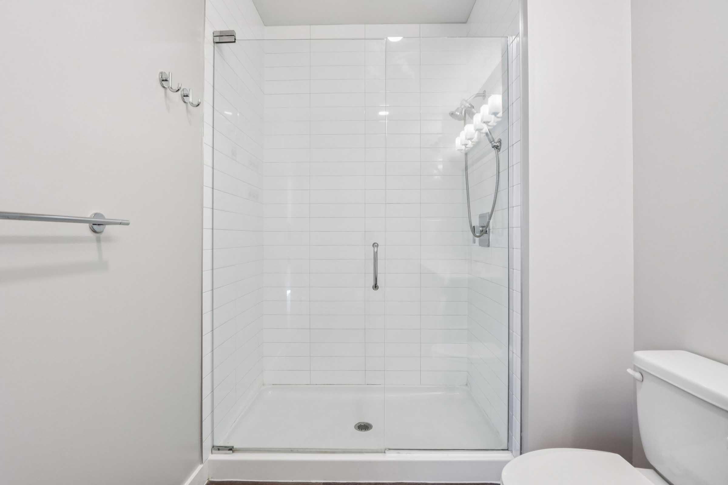 A clean, modern bathroom featuring a glass shower enclosure with white tiled walls, a toilet, and a simple mirror. The lighting is bright, and the space is uncluttered, creating a fresh and spacious look.