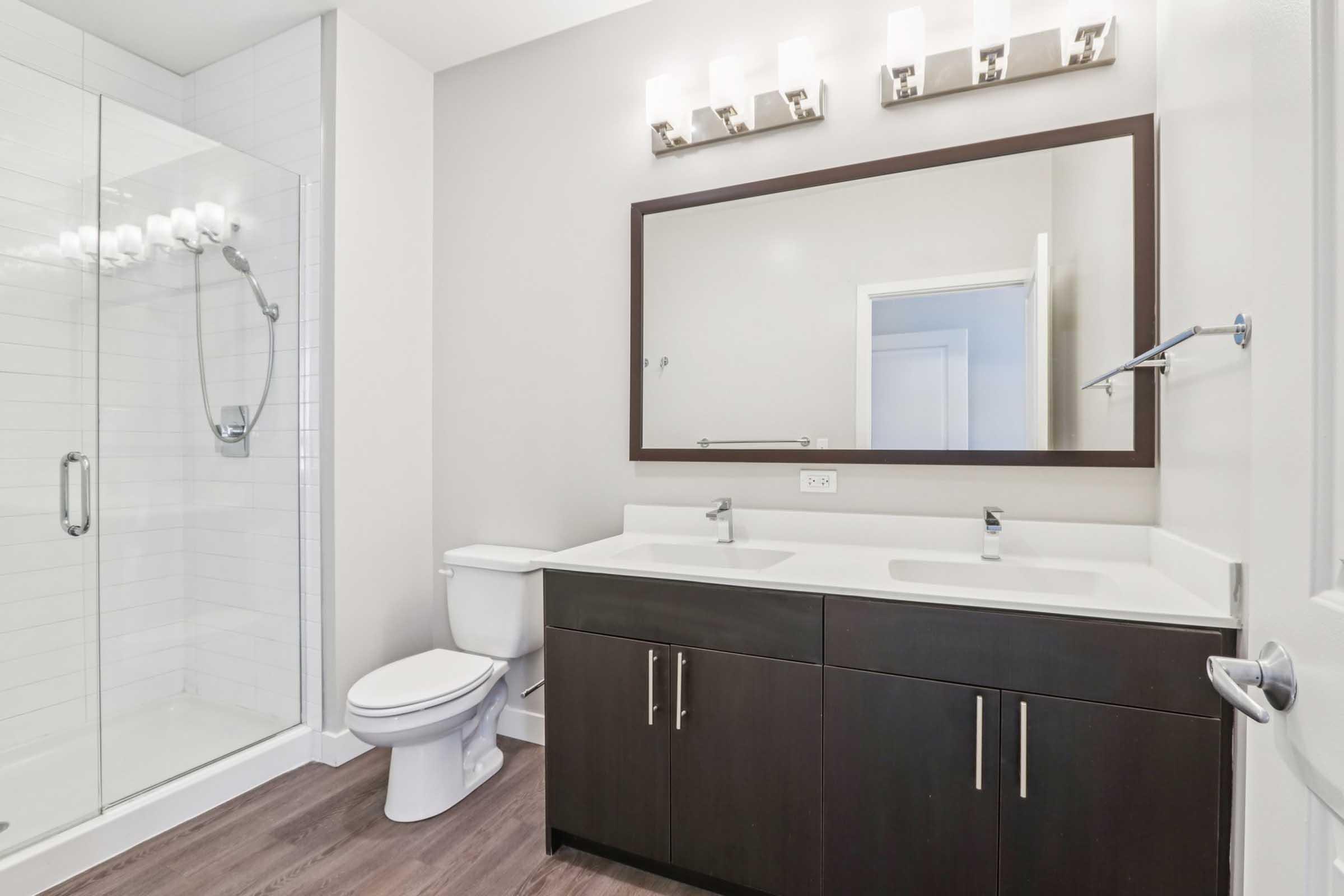 A modern bathroom featuring a glass shower with a white tiled wall, a large mirror above a dual sink vanity with dark wood cabinetry, a white toilet, and stylish light fixtures. The walls are painted in a soft gray, creating a bright and inviting space.