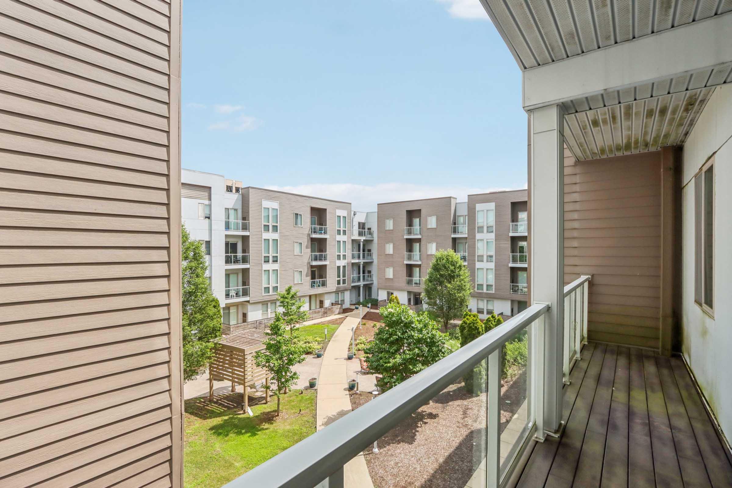 View from a balcony overlooking a landscaped courtyard with modern apartment buildings. The area features green trees, a walking path, and outdoor seating, creating a spacious, inviting environment. The sky is clear, and the architecture is contemporary.