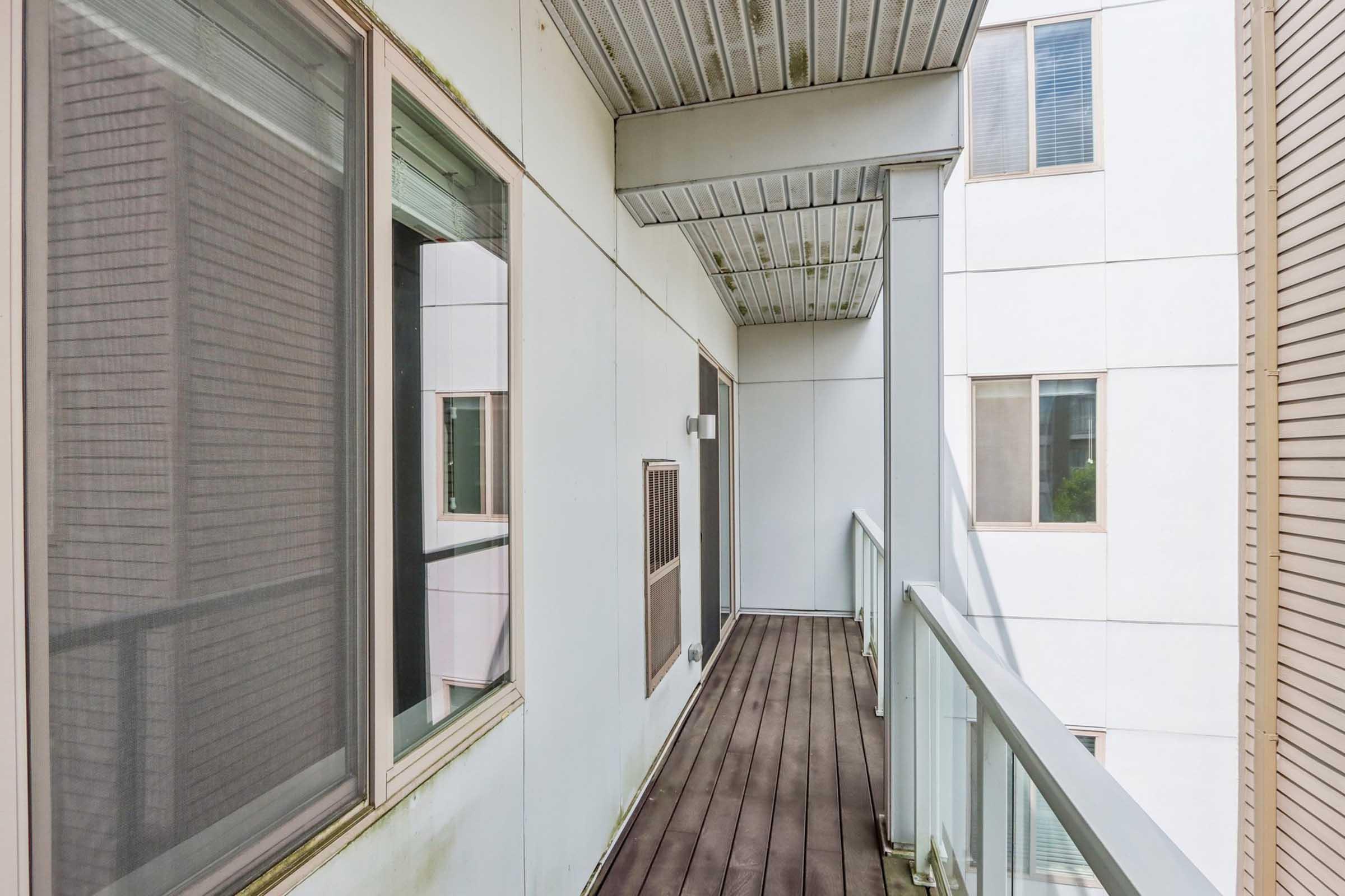 A narrow balcony with wooden flooring and glass railing. The wall is largely white with a few windows reflecting the exterior. There are some signs of wear, such as stains on the walls and ceiling. The setting appears to be part of a modern building.