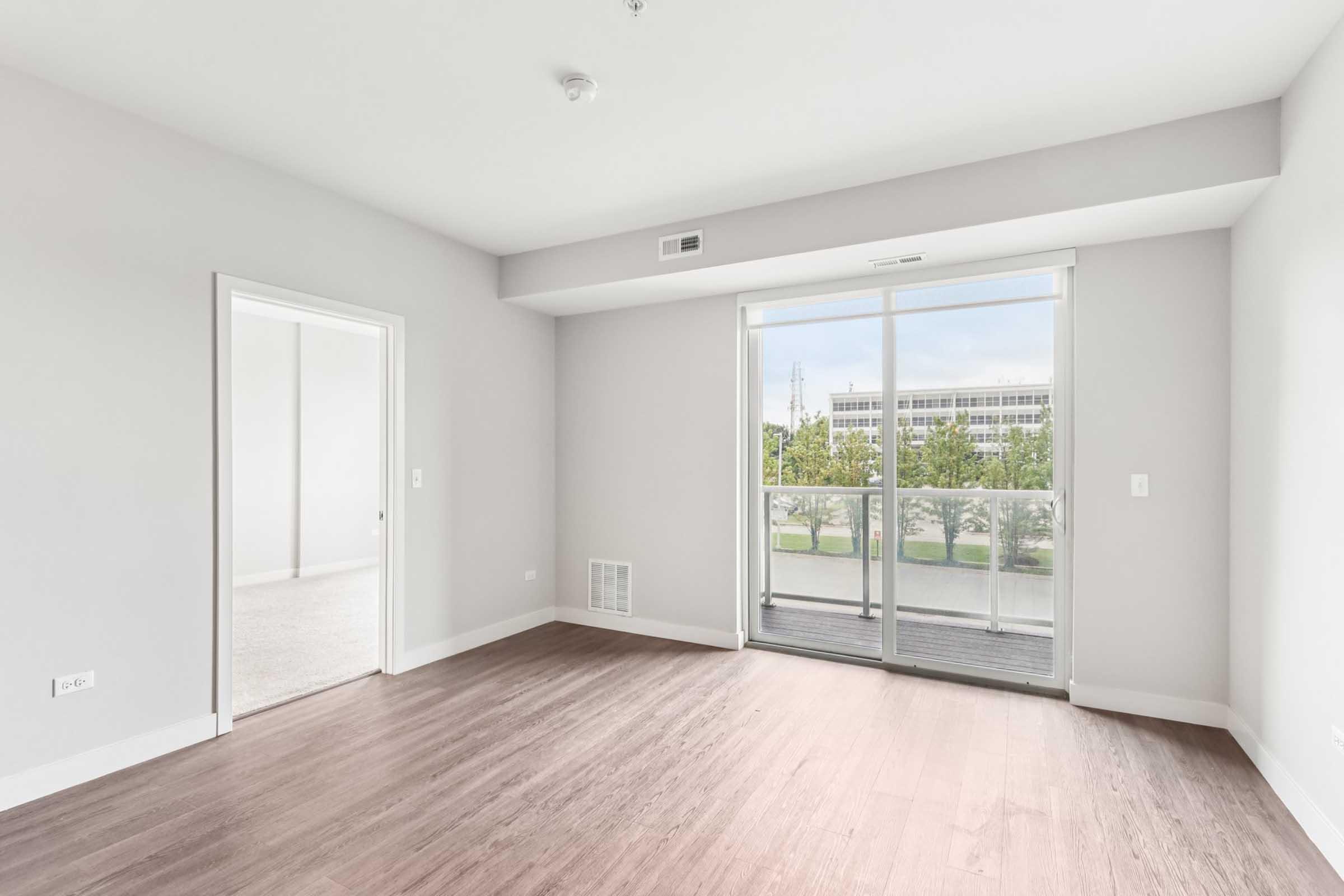 A spacious, empty room with light gray walls and wooden flooring. It features a large window with a balcony door, offering a view of greenery outside. A doorway leads to a second room, and the overall ambiance is bright and modern, suitable for various interior designs.