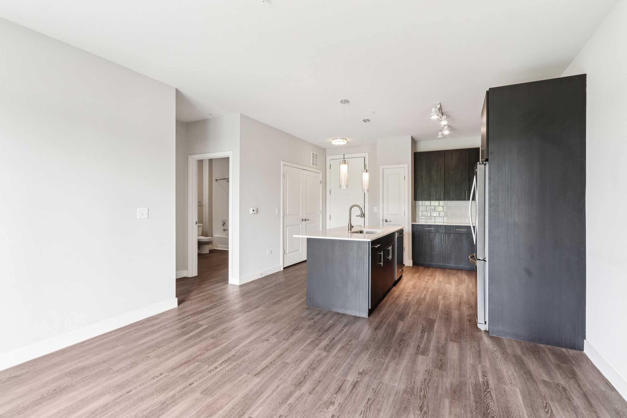 Modern kitchen and living space featuring a central island with a sink, sleek dark cabinetry, and stainless steel appliances. Light-filled room with hardwood flooring and neutral wall colors. Open layout leading to additional rooms, including a bathroom area visible in the background.