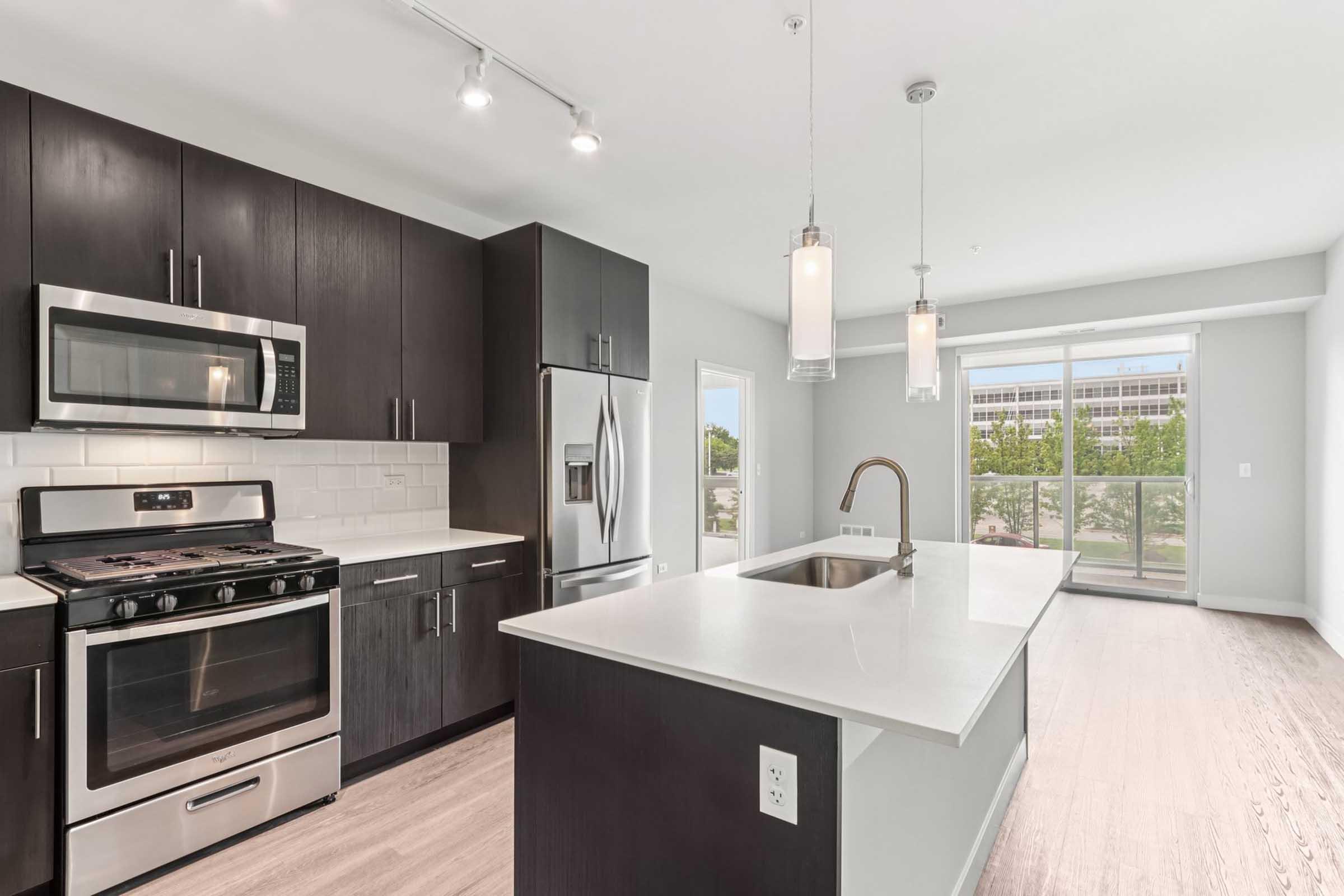 Modern kitchen with dark wood cabinetry, stainless steel appliances, and a large island with a sink. Features include a gas stove, microwave, and a spacious window allowing natural light. The design is contemporary and open, ideal for cooking and entertaining.