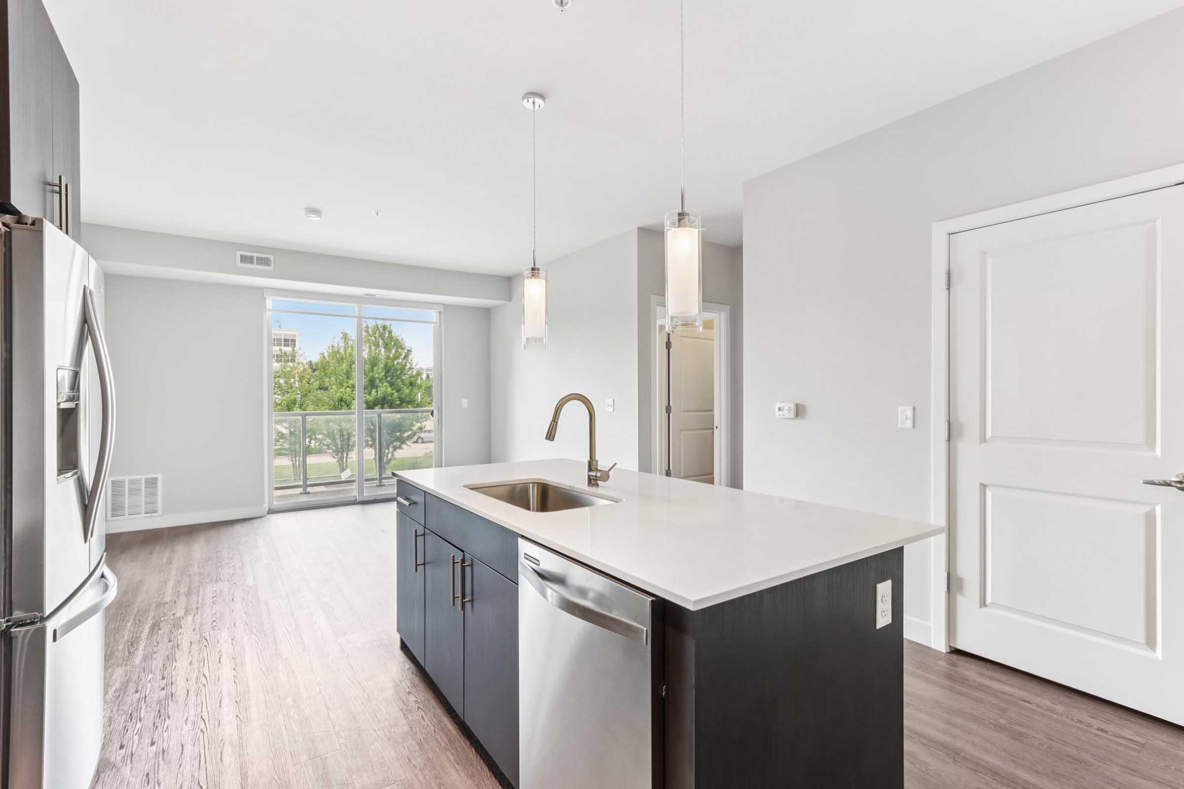 Modern kitchen showcasing stainless steel appliances, including a refrigerator and dishwasher, with a spacious island featuring a sink. Light gray walls and hardwood-like flooring create a contemporary aesthetic. Large windows provide natural light and a view of greenery outside.