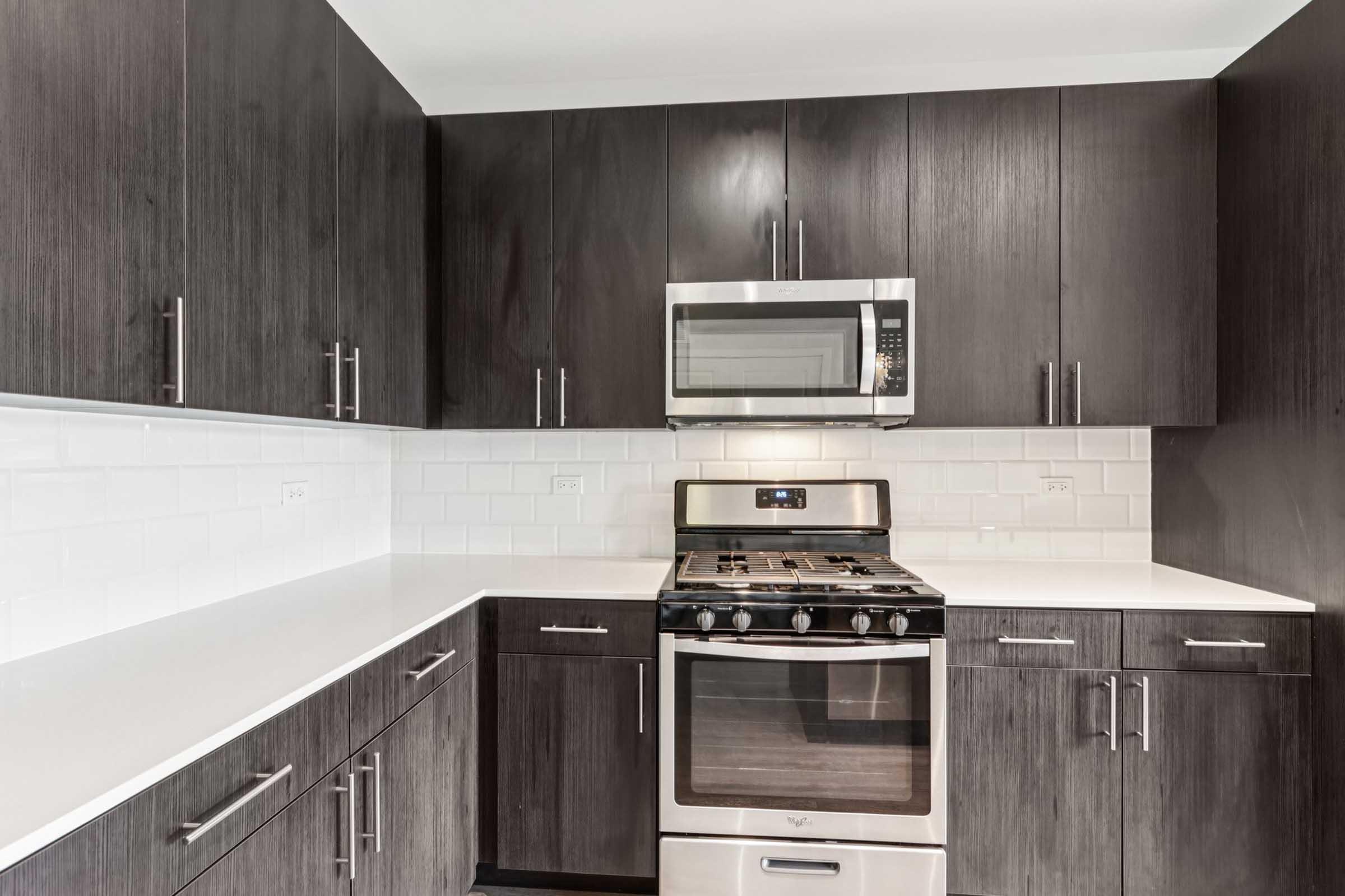 Modern kitchen featuring dark wood cabinets, a stainless steel gas stove, a microwave above it, and a white countertop. The backsplash consists of white subway tiles, creating a clean and contemporary look. The kitchen layout is compact, with ample cabinet space and modern appliances.