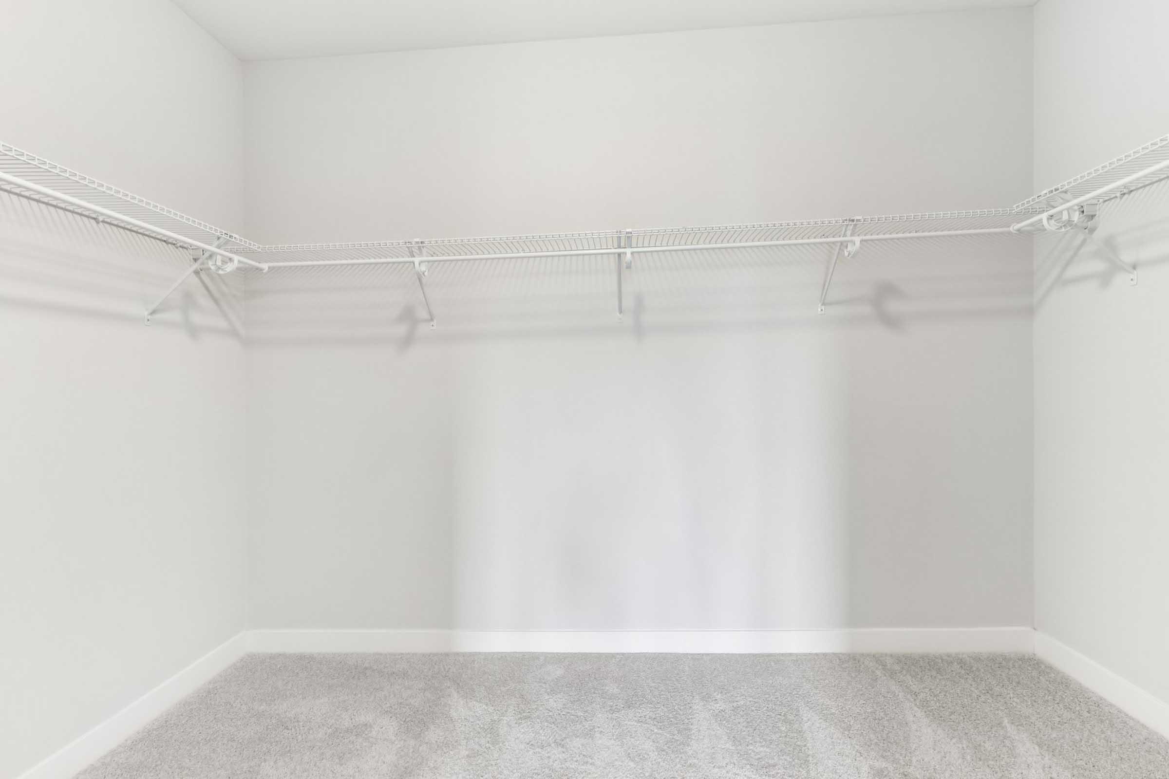 Empty closet with white walls and light gray carpet. Metal wire shelving mounted on the walls, casting soft shadows. The space is well-lit and free of any items, giving a clean and organized appearance.