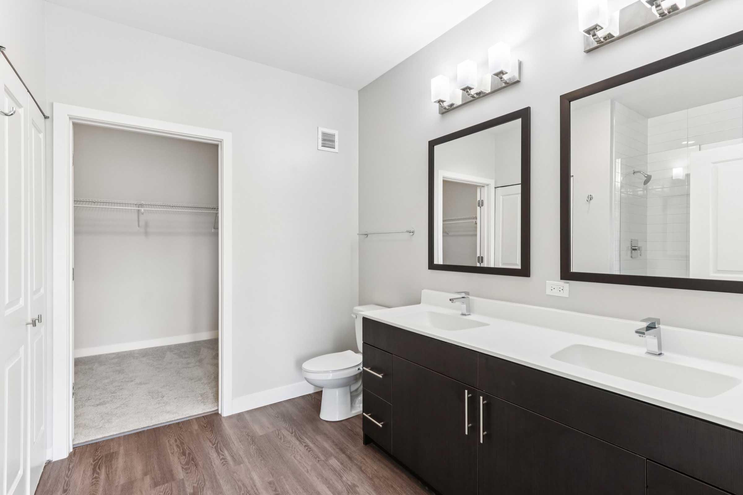 A modern bathroom featuring a double sink vanity with dark cabinetry, large mirrors above the sinks, and stylish wall lighting. There is a separate area with a toilet and a spacious walk-in closet with shelving visible. The walls are painted light gray, and the flooring is a warm wood tone.