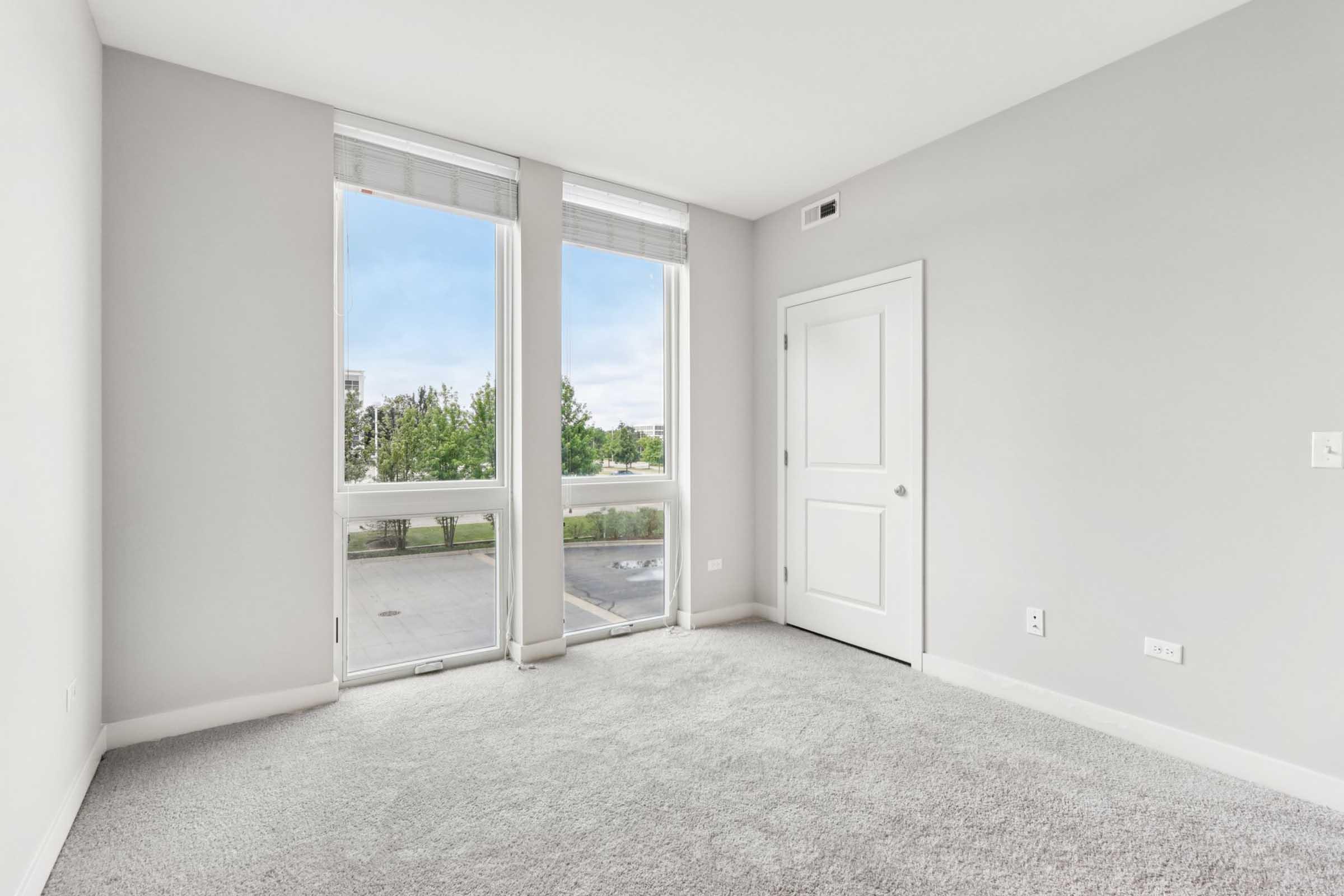 A bright, empty room with light gray walls and plush carpeting. Large windows allow natural light to flood in, providing a view of greenery outside. A white door is visible, adding to the modern aesthetic of the space. The overall atmosphere is clean and inviting, ideal for personalization.