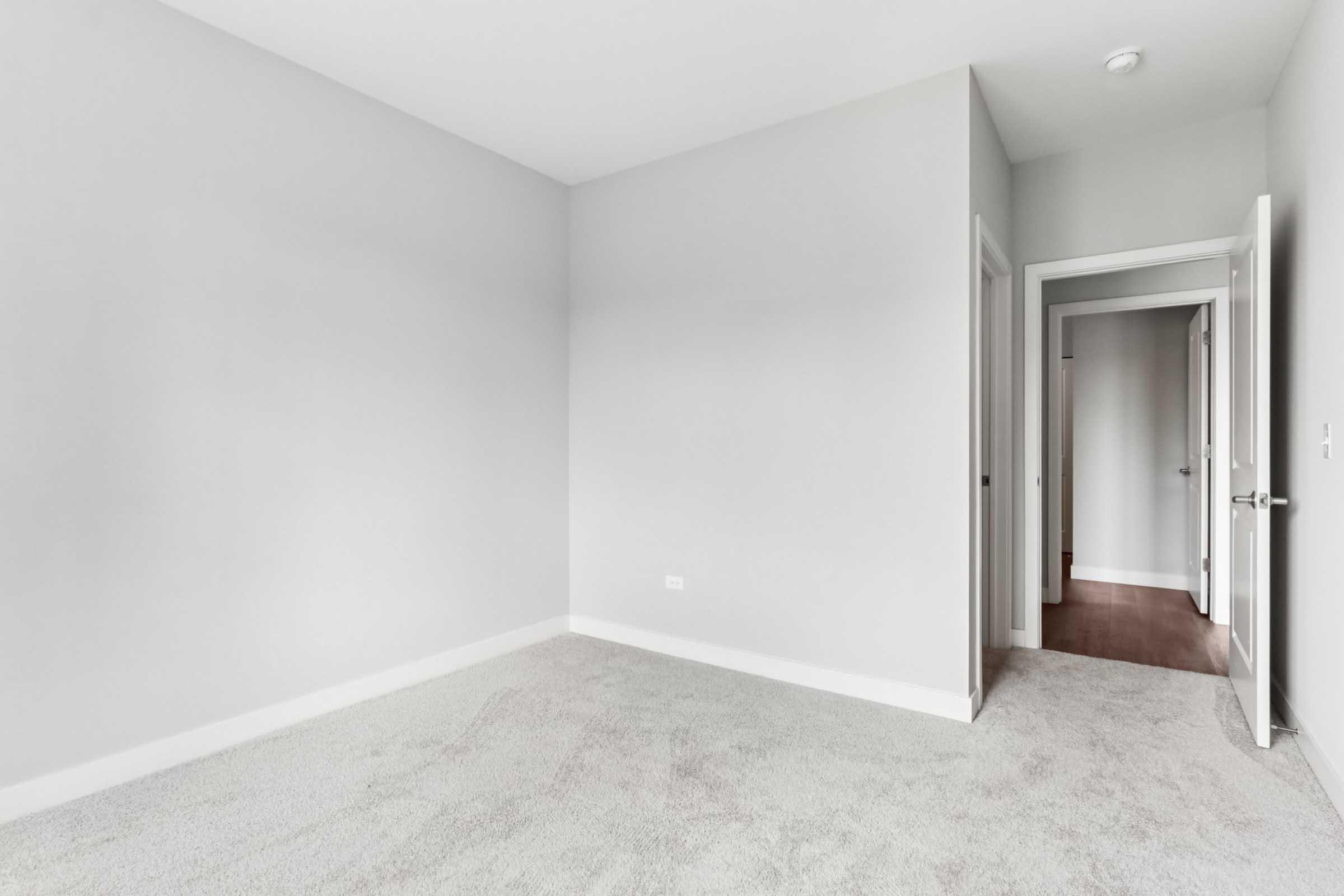 An empty room with light gray walls and soft, beige carpet. The room features a doorway on the right leading to a hallway, and there is a door slightly ajar in the background. The space is well-lit with natural light.