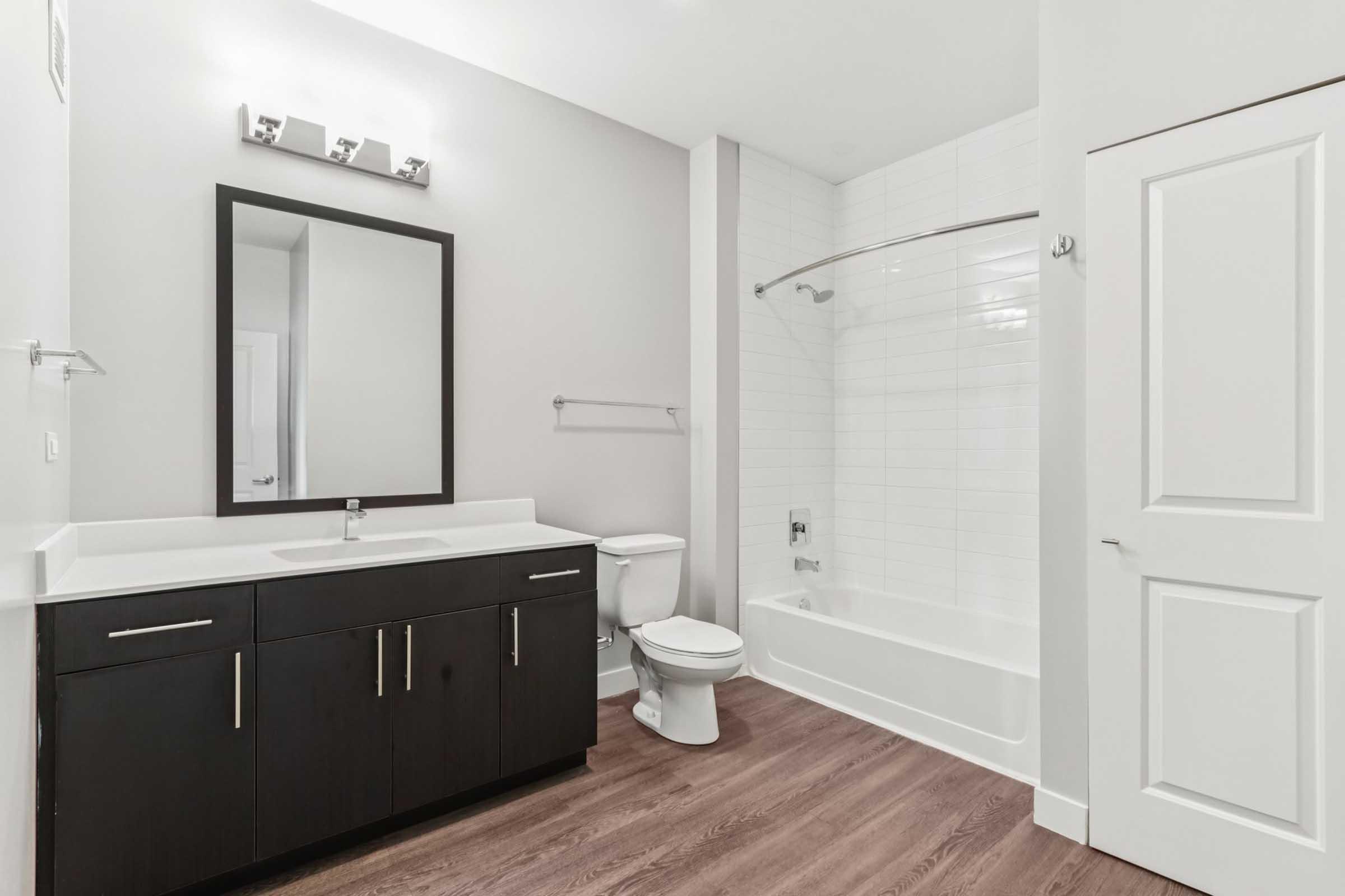 Modern bathroom featuring a white tub with a curved glass shower rod, a dark wood vanity with a sink, a large mirror above the vanity, a toilet, and a light-colored wall. The flooring is wooden, and there is a door leading to another room. The overall design is clean and contemporary.