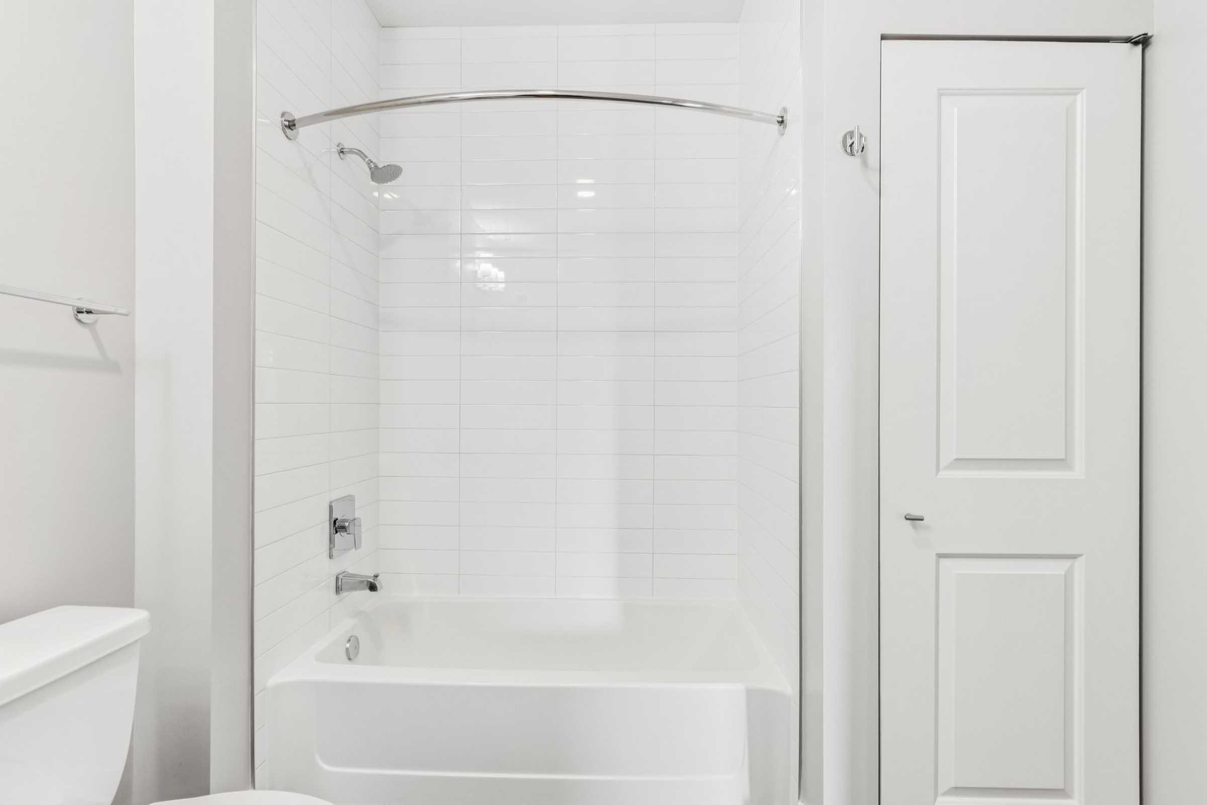 A clean, modern bathroom featuring a white bathtub with a shower curtain rod, tiled walls, and a white toilet. The room is well-lit and has a minimalist design, with a door visible on the right side.