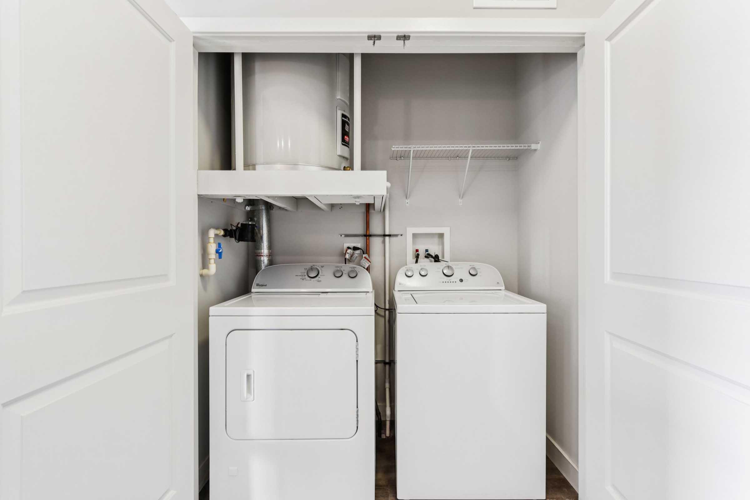 A laundry room featuring a white washing machine and dryer side by side, with a water heater mounted above. The room has light-colored walls and a shelf for storage above the appliances. Two white doors are slightly open, revealing the appliances inside a compact space.