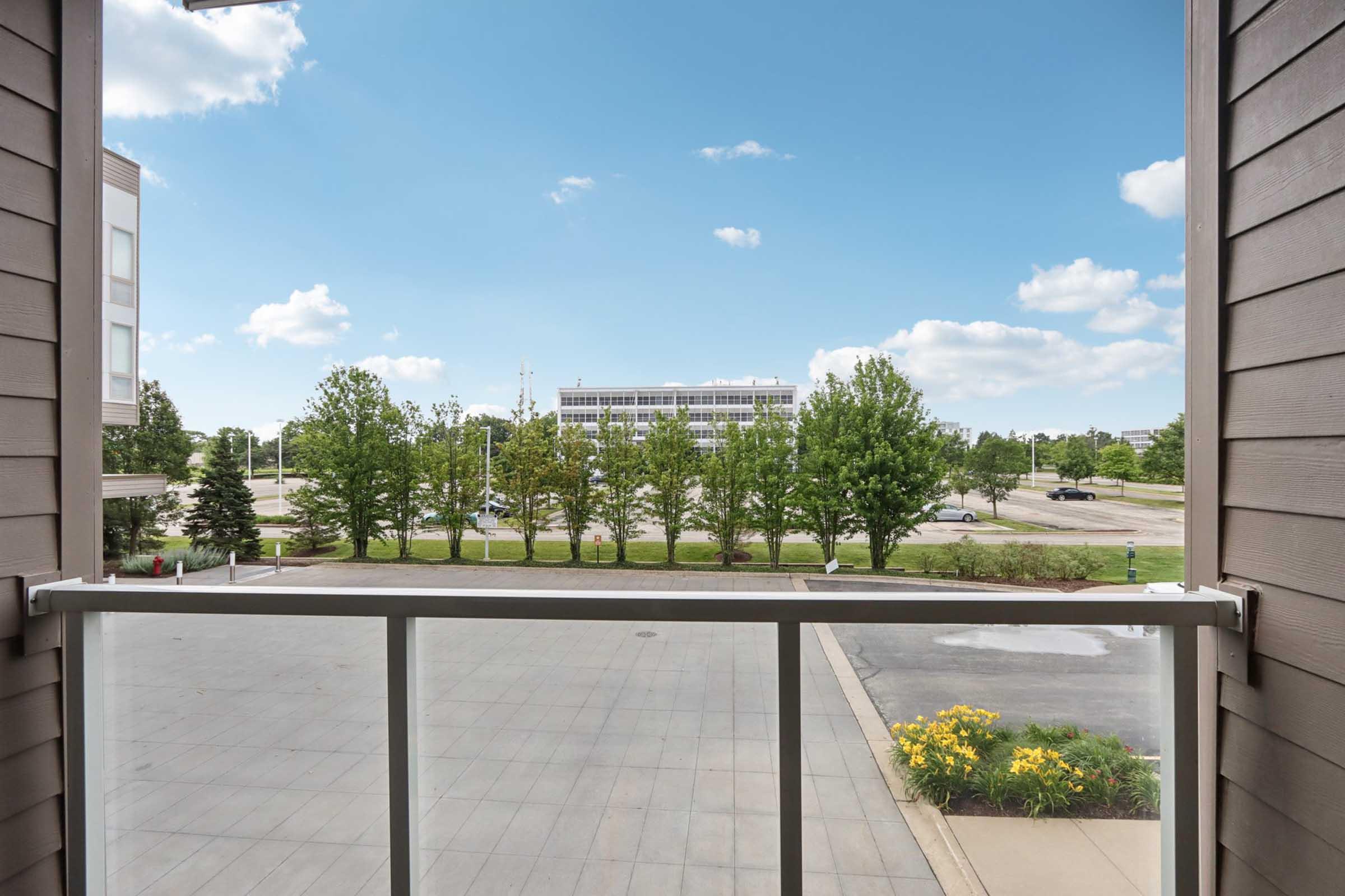 View from a balcony overlooking a parking lot and a row of trees under a clear blue sky. The scene includes a modern building in the background and colorful flowers in the foreground, creating a pleasant and inviting atmosphere.