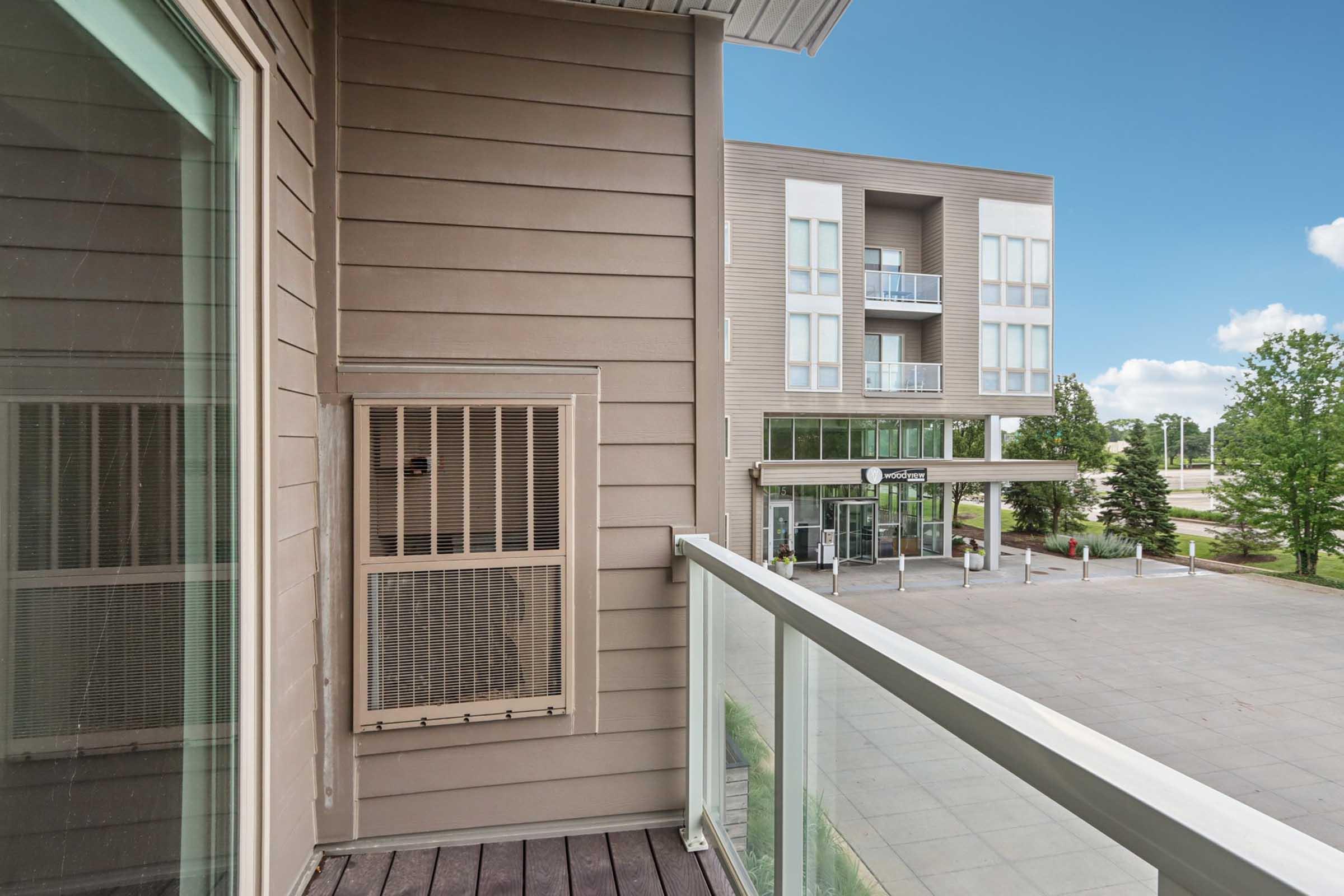 View from a balcony showing a modern building in the background with large windows and a welcoming entrance. An air conditioning unit is mounted on the wall of the balcony, and the scene includes a clear blue sky and greenery in the vicinity.