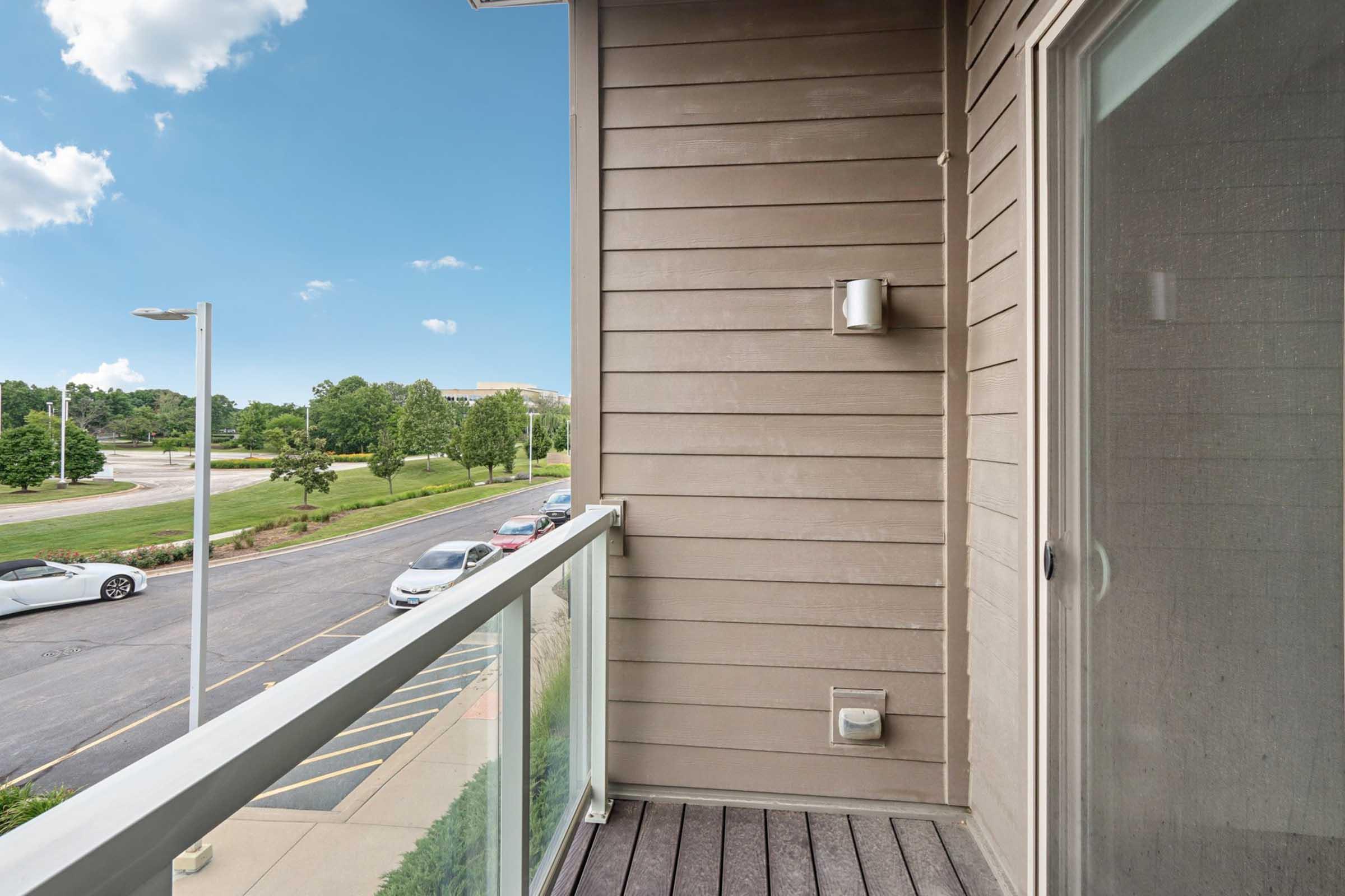 A small balcony featuring wooden flooring and a glass railing, overlooking a tree-lined street. There are several parked cars visible in the foreground, and a clear blue sky with a few clouds in the background.