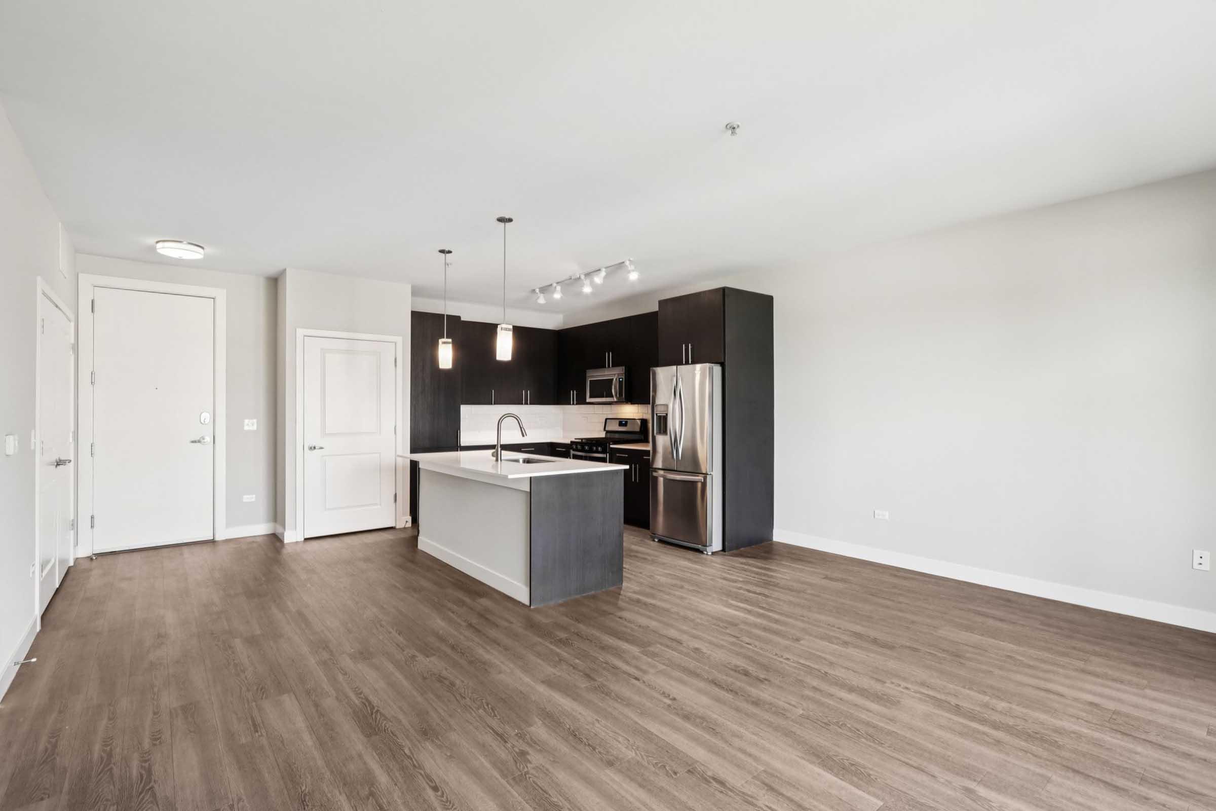 Modern kitchen and living space featuring light wood flooring, dark cabinetry, stainless steel appliances, and a central island with bar seating. Bright natural light fills the area, enhancing the open layout and minimalist design. Suitable for contemporary living.
