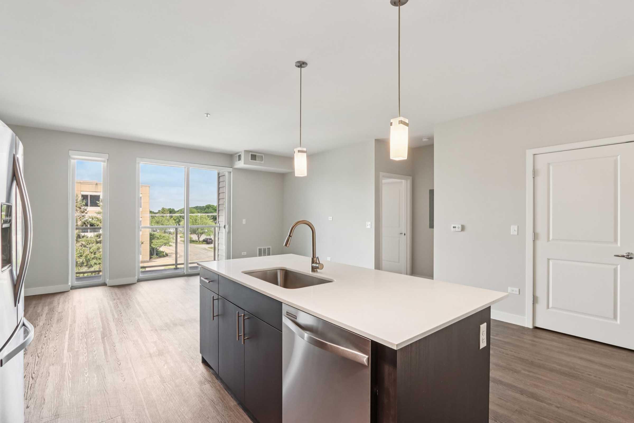 Modern kitchen with an island, featuring stainless steel appliances, pendant lighting, and a sink. Large windows provide natural light and a view of greenery outside. The space has a contemporary design with light-colored walls and hardwood flooring.