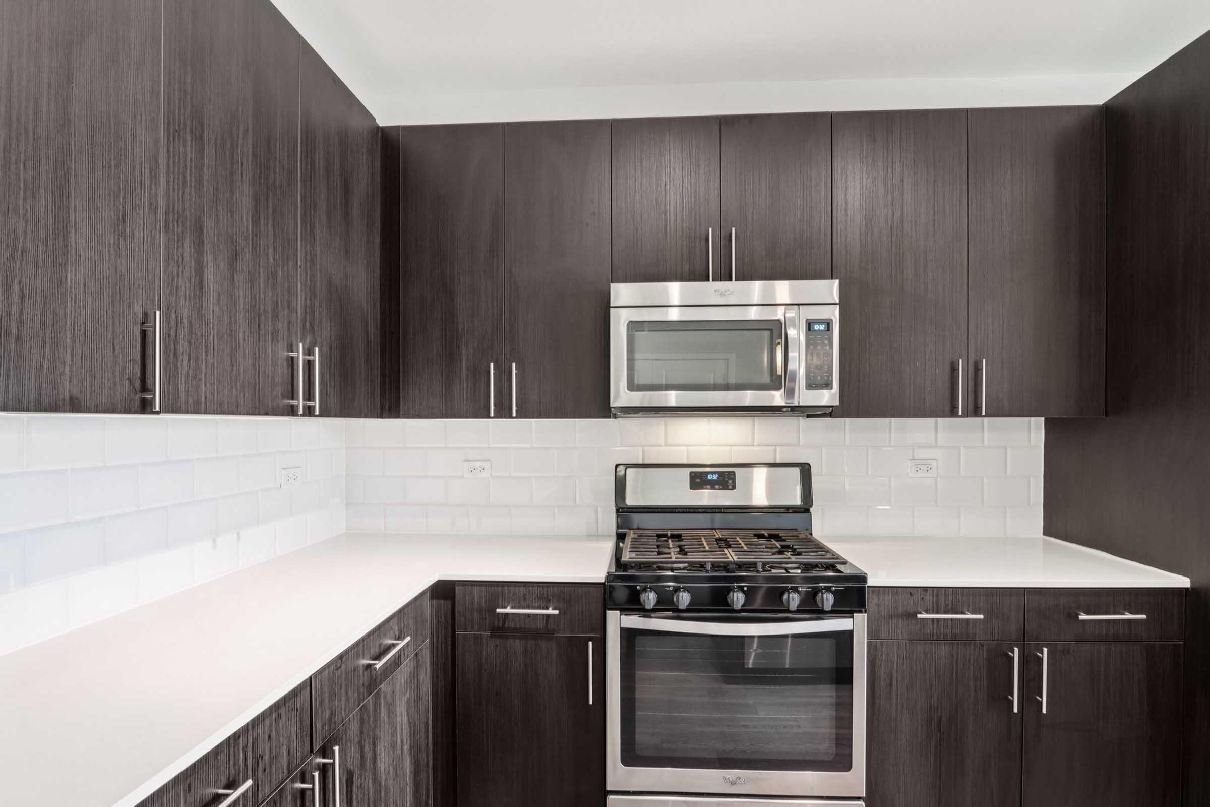 Modern kitchen featuring dark wood cabinetry, stainless steel appliances, including a gas stove, microwave, and a white countertop. The backsplash consists of white subway tiles, creating a sleek and contemporary look. Ample storage space is evident with stylish cabinet handles.