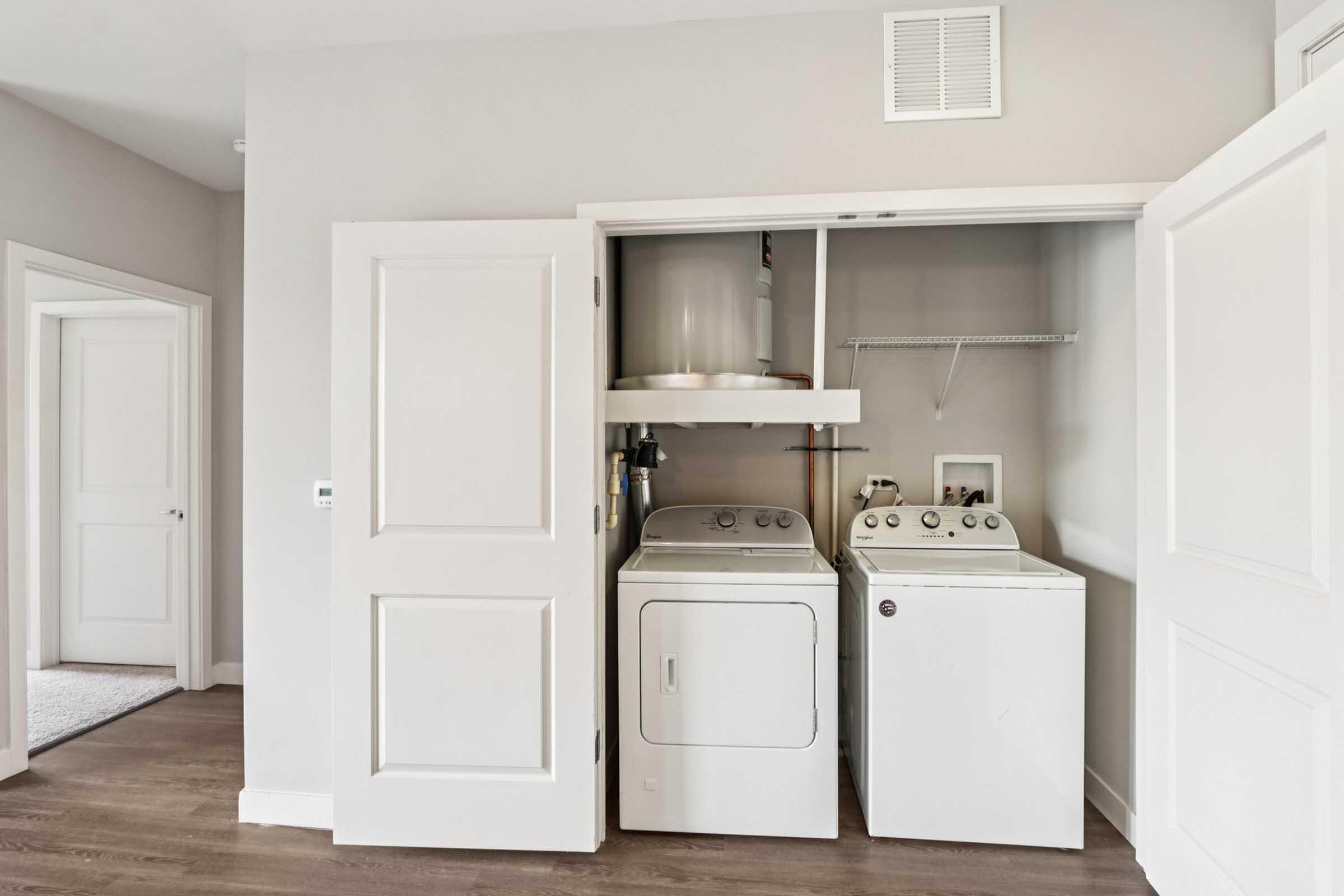 A small laundry area featuring a stacked washer and dryer inside a closet. The space has light gray walls and wooden flooring, with a door leading to another room visible in the background.