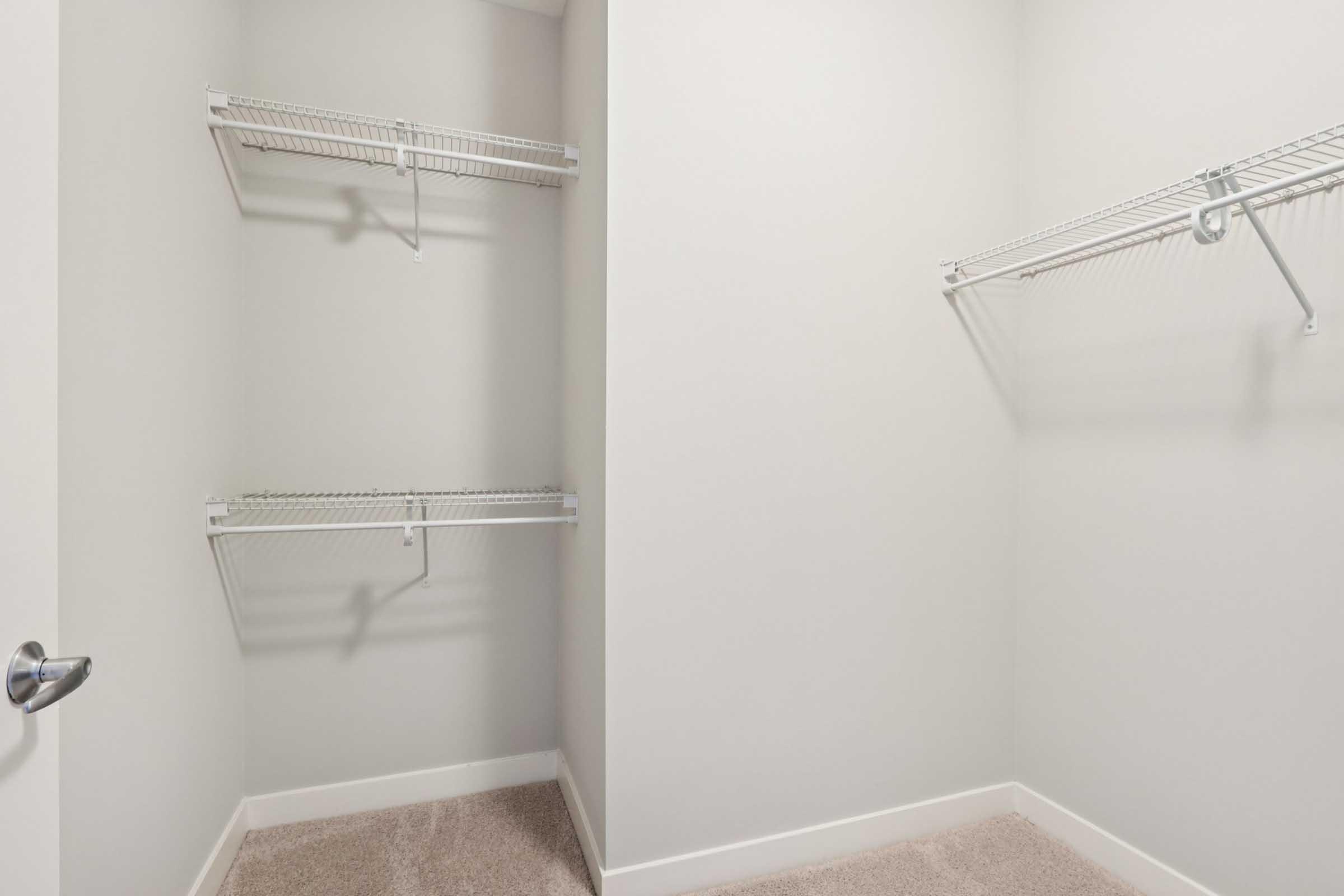 Empty closet with light gray walls and beige carpet. Two white wire shelves are attached to the walls on the left, while the right wall remains bare. The closet door is slightly ajar, revealing the clean, minimalistic interior.