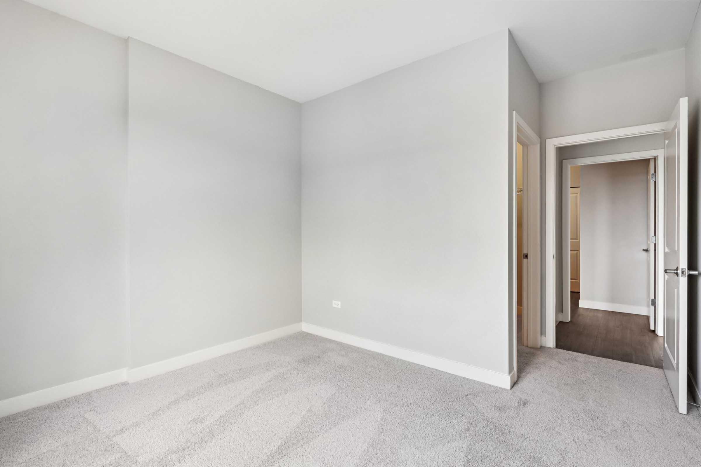 A blank, empty room with light gray walls and carpet. There's a door on the right leading to another area, and the space is well-lit, giving a feeling of openness. The floor has a clean, modern look, suitable for various interior design options.