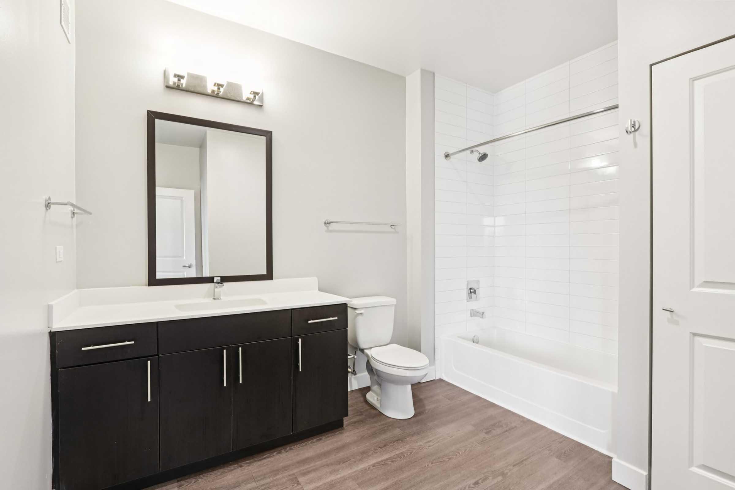Modern bathroom featuring a sleek design with a light gray wall, a dark wood vanity with a white sink, a full-size bathtub with a shower, and a large mirror. The flooring is a warm wood finish, and there is a door visible that leads to another space.