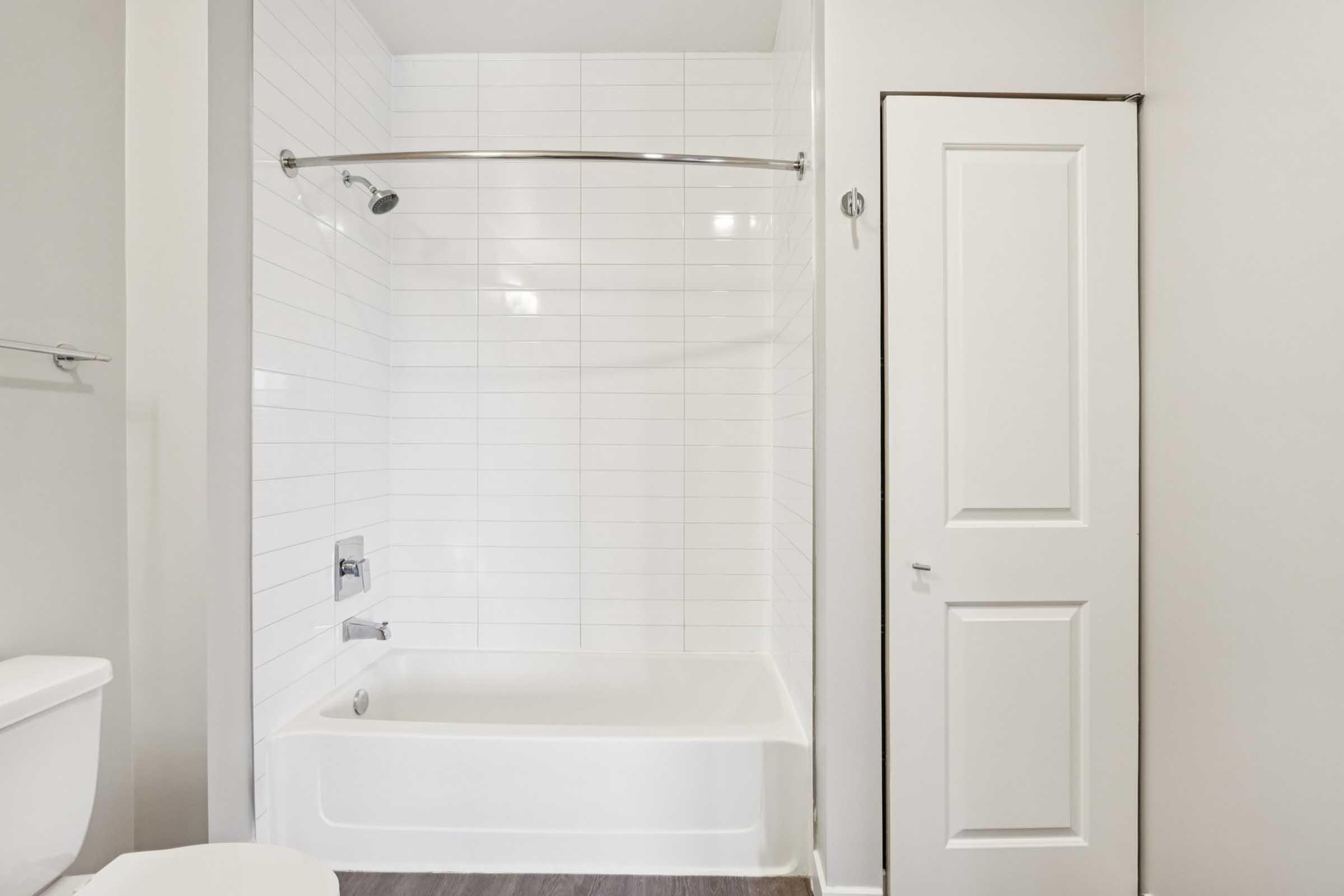 A clean bathroom featuring a white bathtub with a shower rod and curtain, a modern toilet, and a door leading to another area. The walls are tiled in white, and the flooring appears to be a light-colored laminate or vinyl. The space is well-lit and minimalist in design.