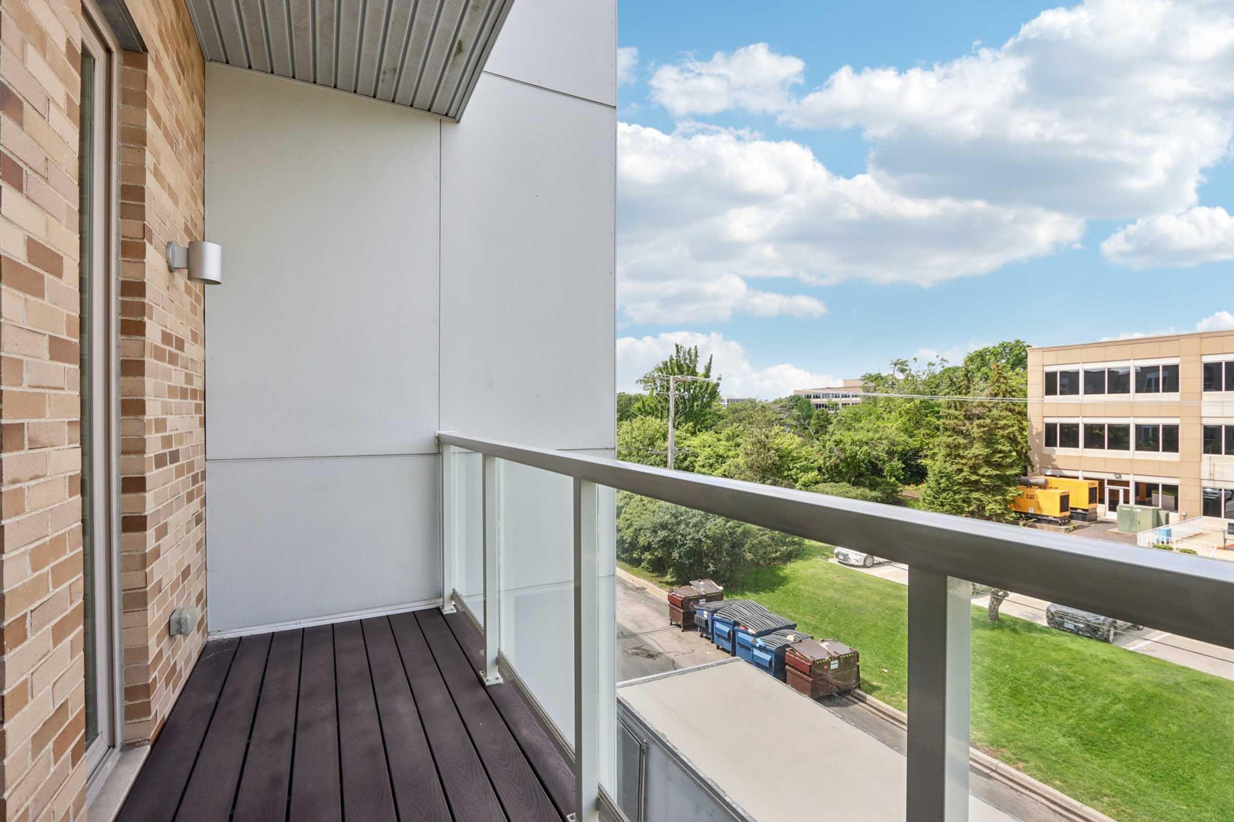 A spacious balcony with a glass railing, featuring wooden flooring. The view includes a grassy area and dumpsters along a street, with a building visible in the background under a partly cloudy sky.