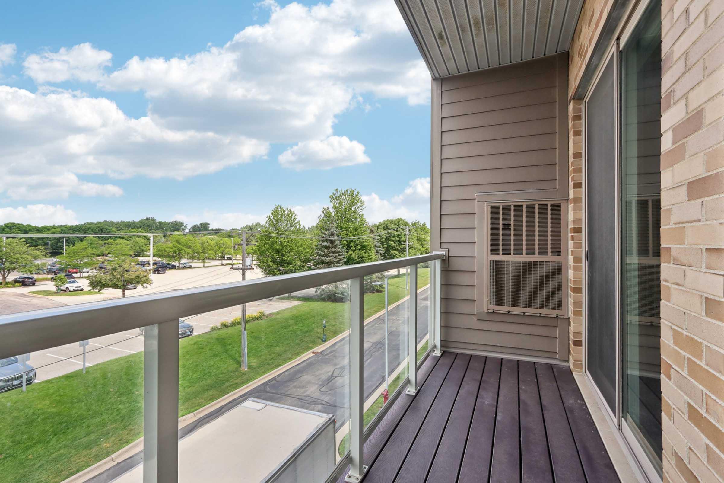 A balcony with wooden flooring and a glass railing, featuring an air conditioning unit. In the background, there are green trees and a parking lot under a partly cloudy sky.