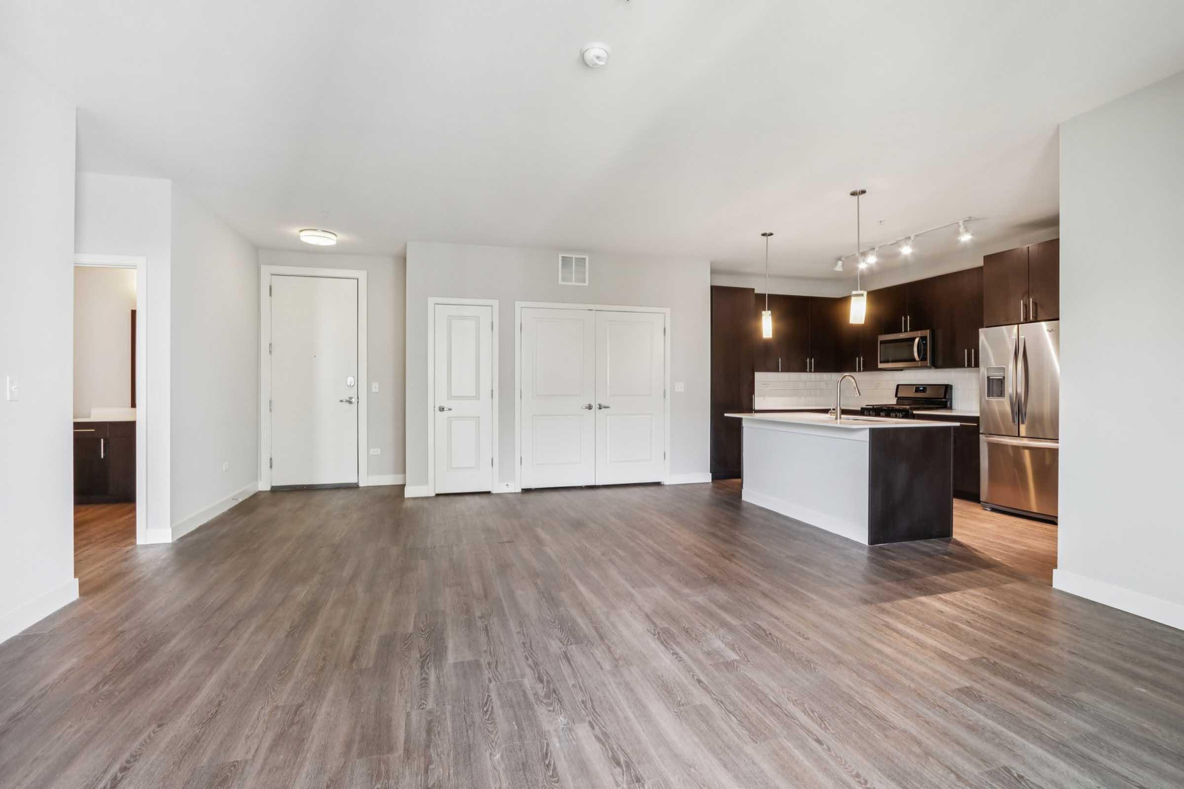 Open-concept living area featuring modern hardwood flooring, with a view of a kitchen equipped with stainless steel appliances and dark cabinetry. Light-colored walls create a bright atmosphere, and there are two doors leading to other areas. Recessed lighting adds to the contemporary design.
