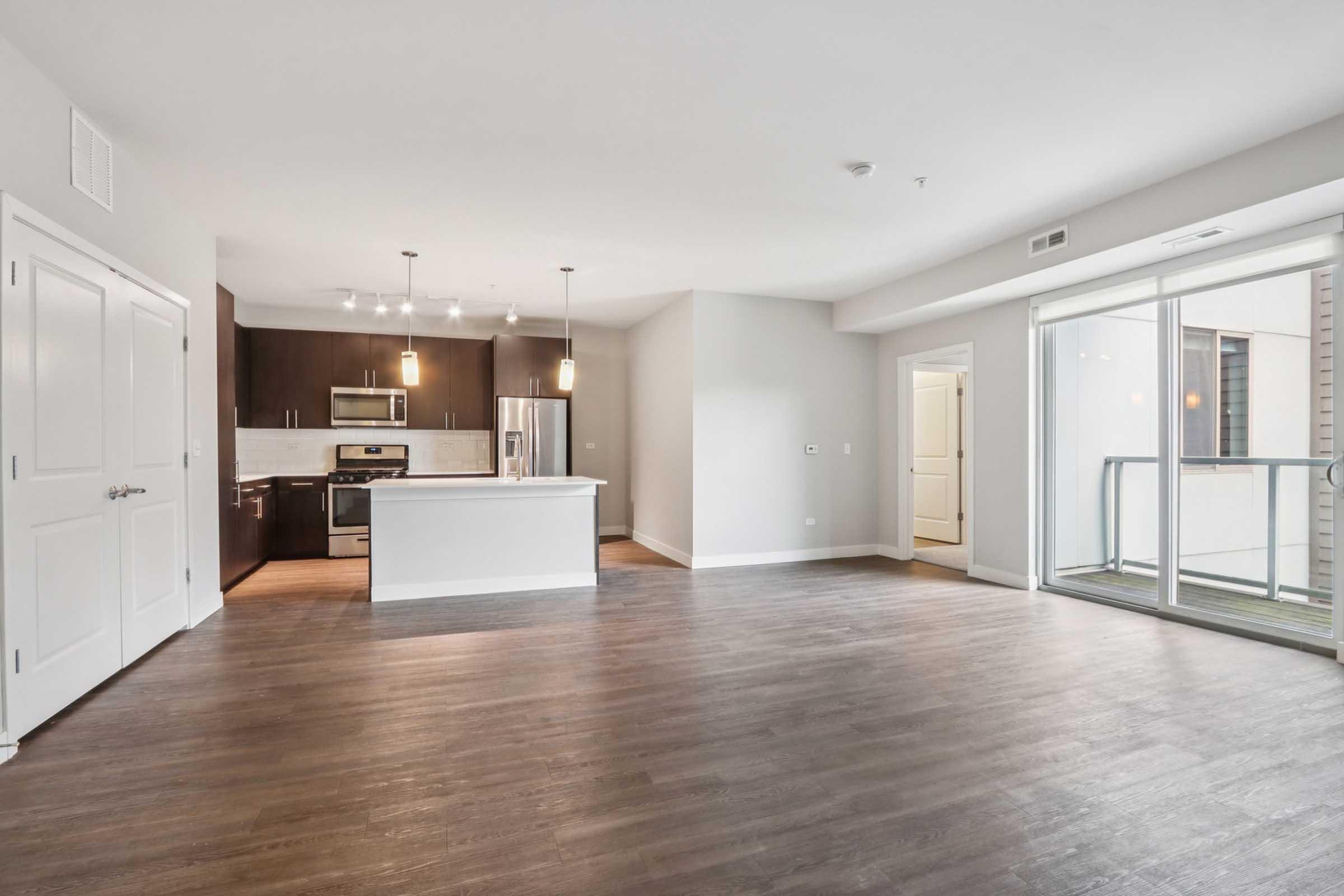 Spacious modern apartment interior featuring an open-concept living area with hardwood floors, a kitchen equipped with stainless steel appliances, and a balcony with sliding glass doors. The design includes neutral wall colors and contemporary lighting fixtures, creating a bright and inviting atmosphere.