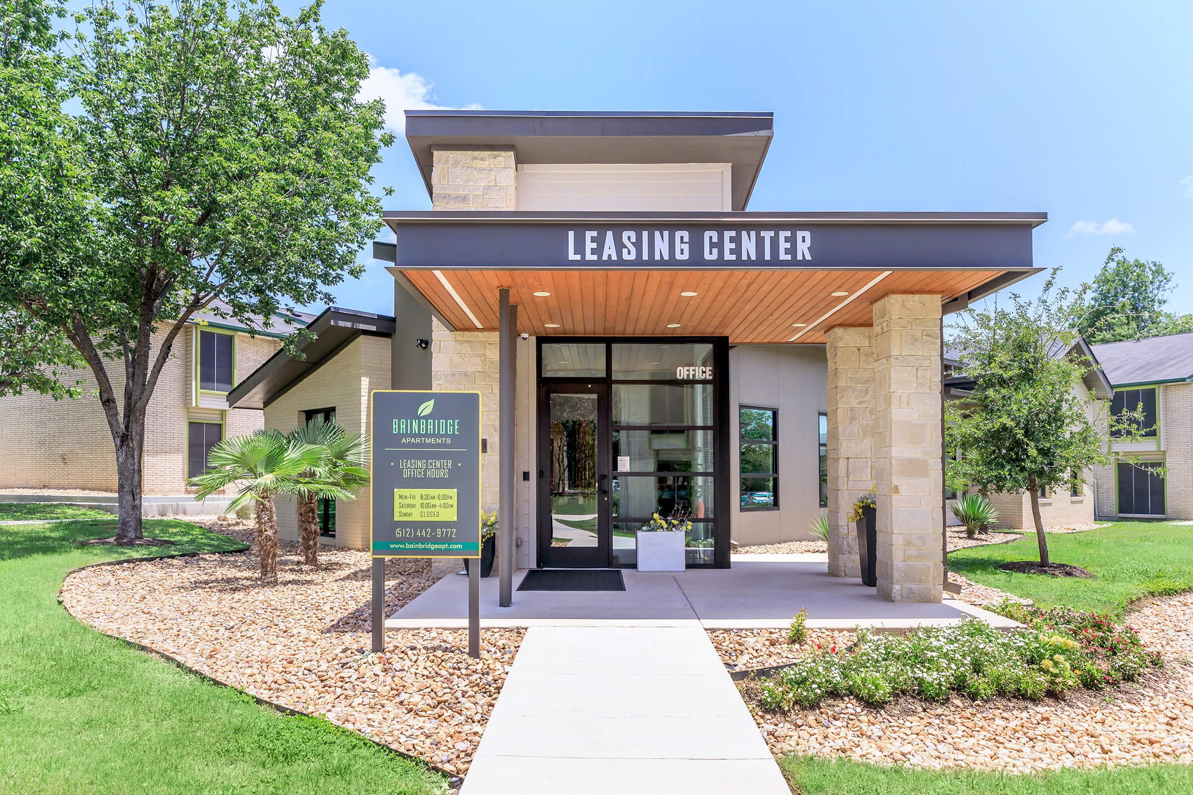 A modern leasing center with a stone and wood exterior, featuring large glass doors. The building is surrounded by lush green grass and landscaping with palm trees. A sign indicates it's the leasing center, with contact information visible. Bright, sunny day with clear skies.