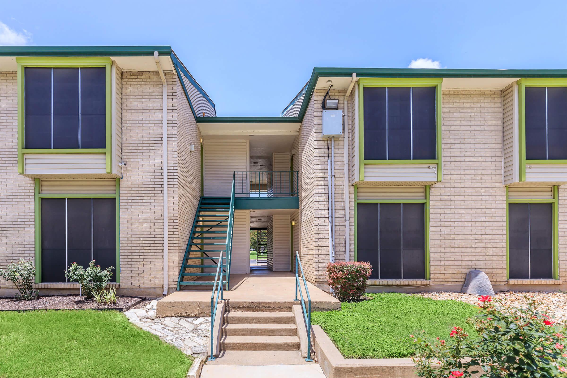 Exterior view of a two-story apartment building featuring green trim, large windows, and a central staircase leading to the upper level. The entrance is flanked by well-maintained shrubs and flowers, with a concrete pathway and a small landscaped area. Bright blue sky overhead adds to the inviting atmosphere.