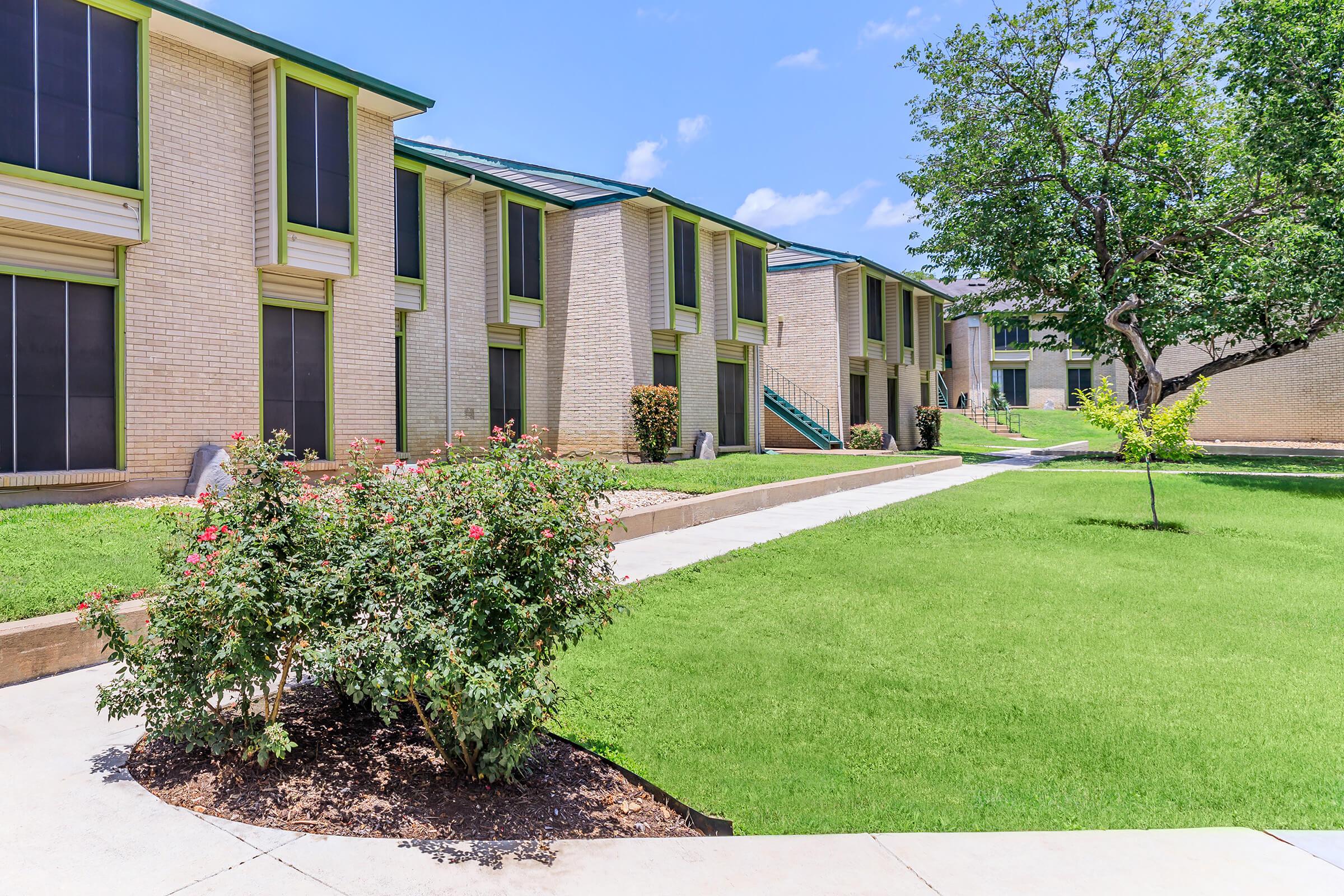 A well-maintained residential complex featuring multiple buildings with light-colored brick exteriors. There are manicured lawns, shrubs, and a small tree. The scene is set under a clear blue sky, showcasing a peaceful and inviting atmosphere in a suburban area.