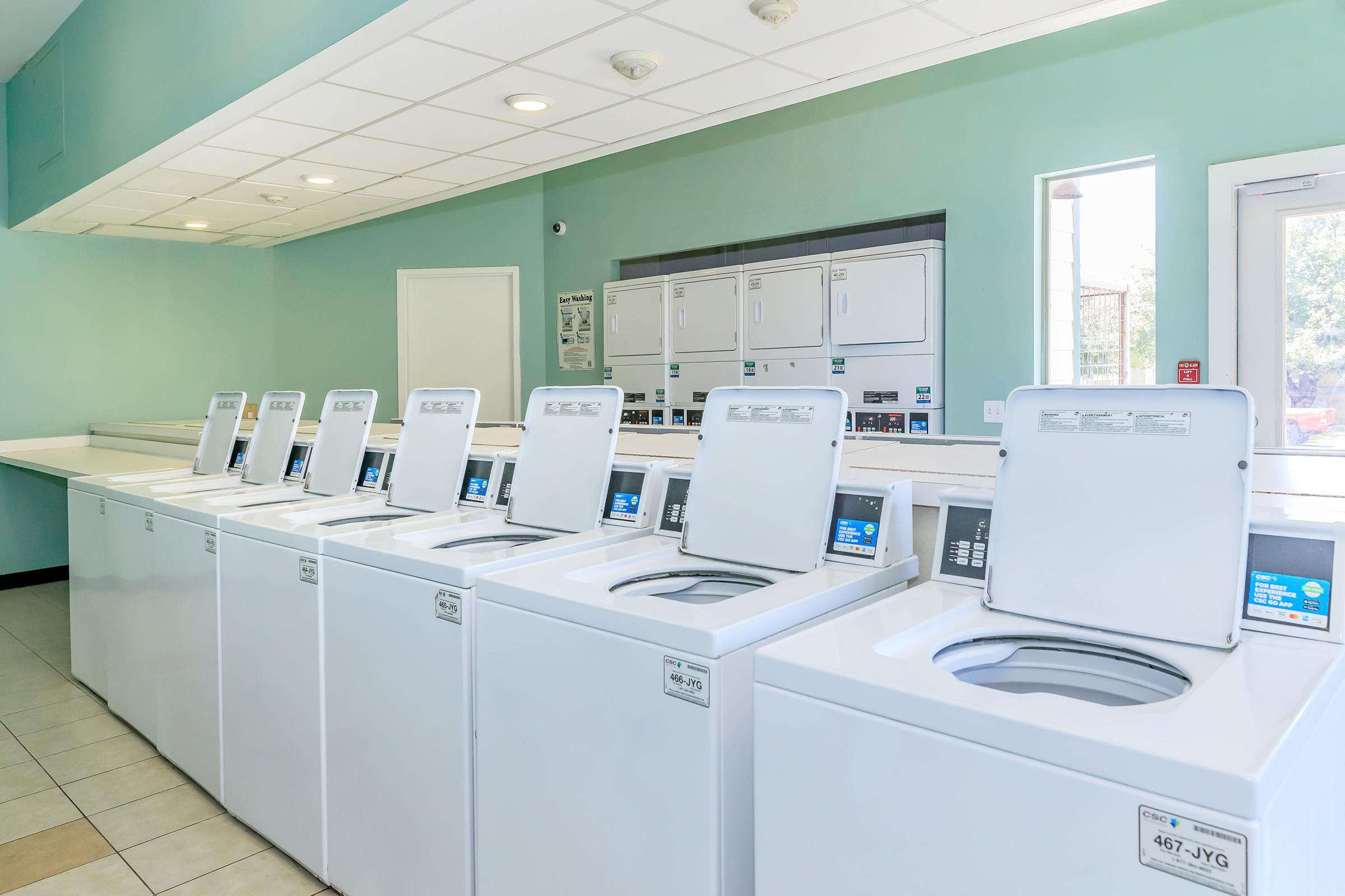 A clean and organized laundry room featuring a row of white washing machines against a light blue wall. Large windows allow natural light to illuminate the space, and there are several drying machines in the background. The floor is tiled, contributing to the modern and well-maintained appearance of the facility.
