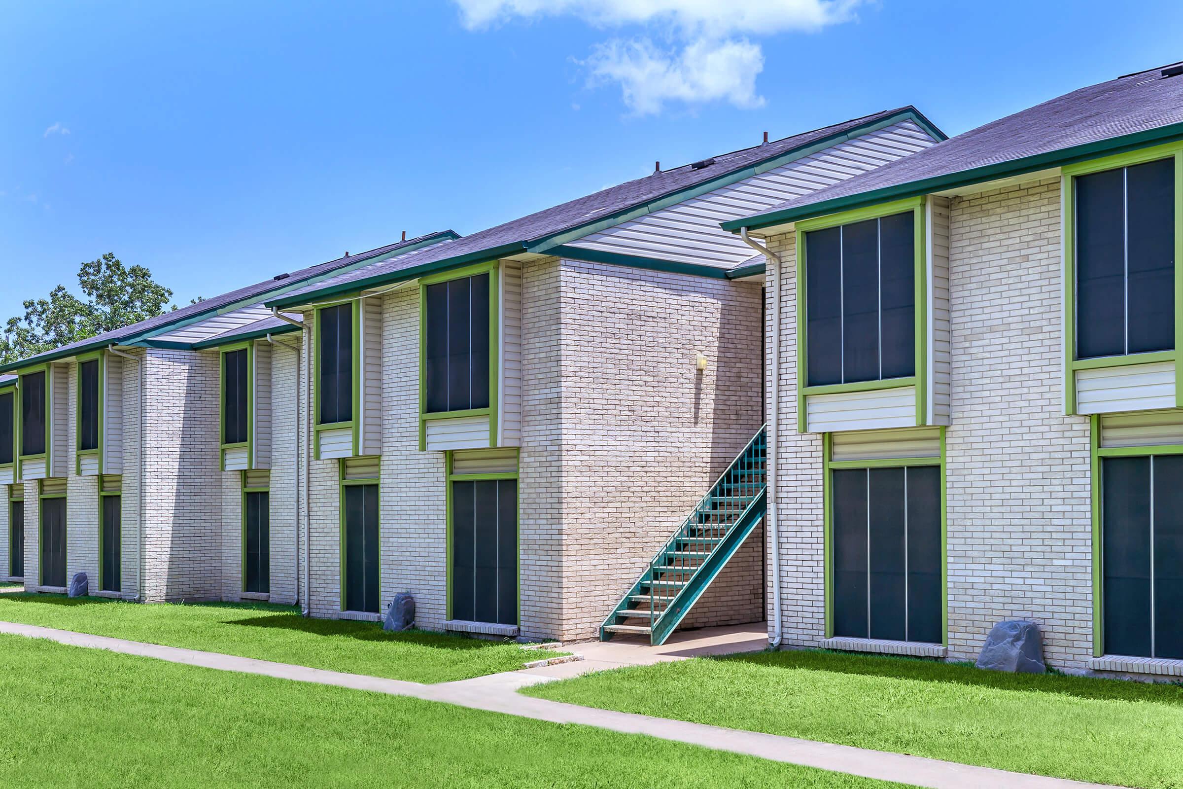 Row of residential buildings with multiple units, featuring large windows and a green staircase leading to the upper level. The buildings have a light brick exterior and are surrounded by well-maintained grass and a clear blue sky.