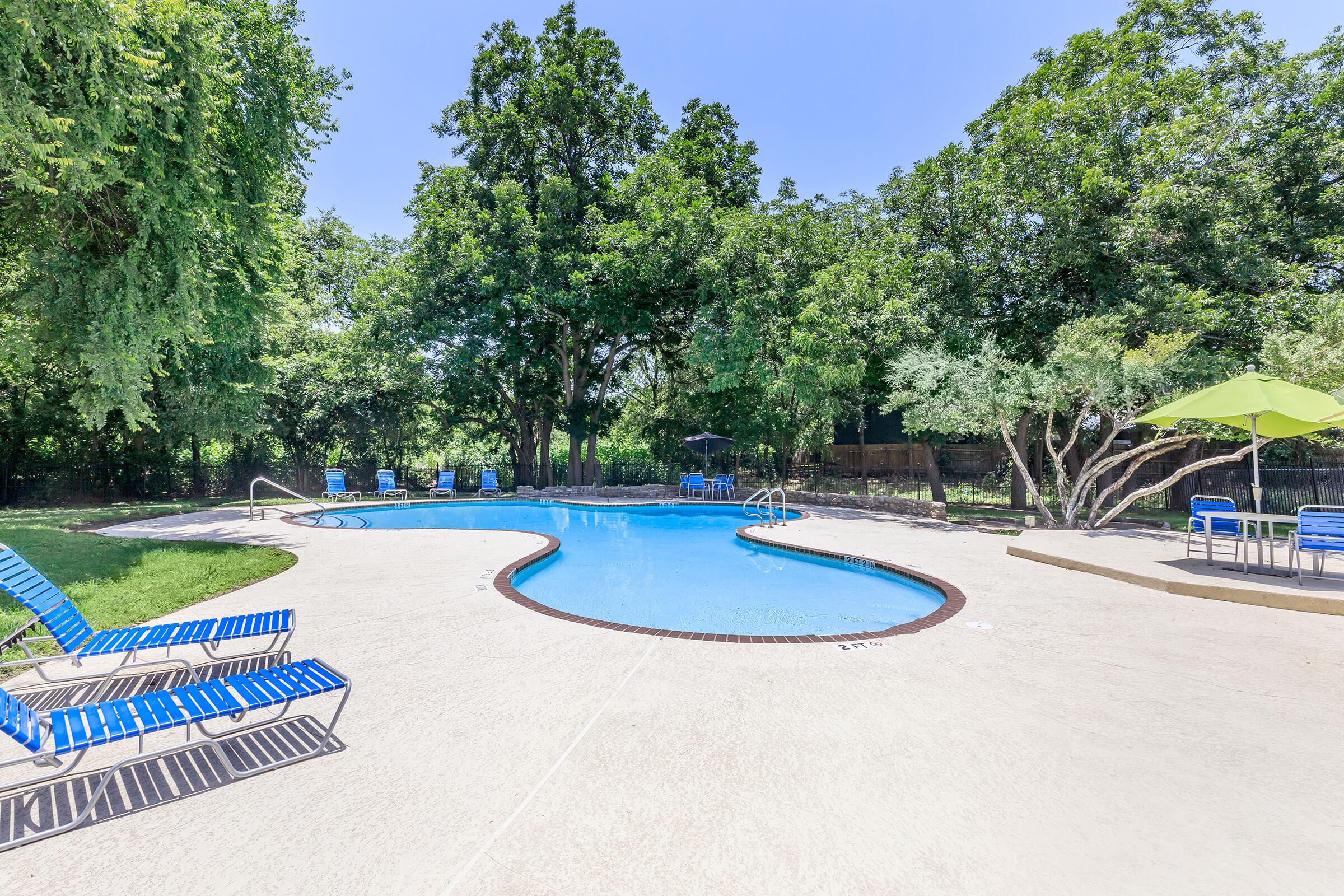 A secluded swimming pool surrounded by lush green trees. Several blue lounge chairs are positioned on the pool deck, and there are umbrellas for shade. The clear blue water reflects the bright blue sky, creating a relaxing outdoor atmosphere.