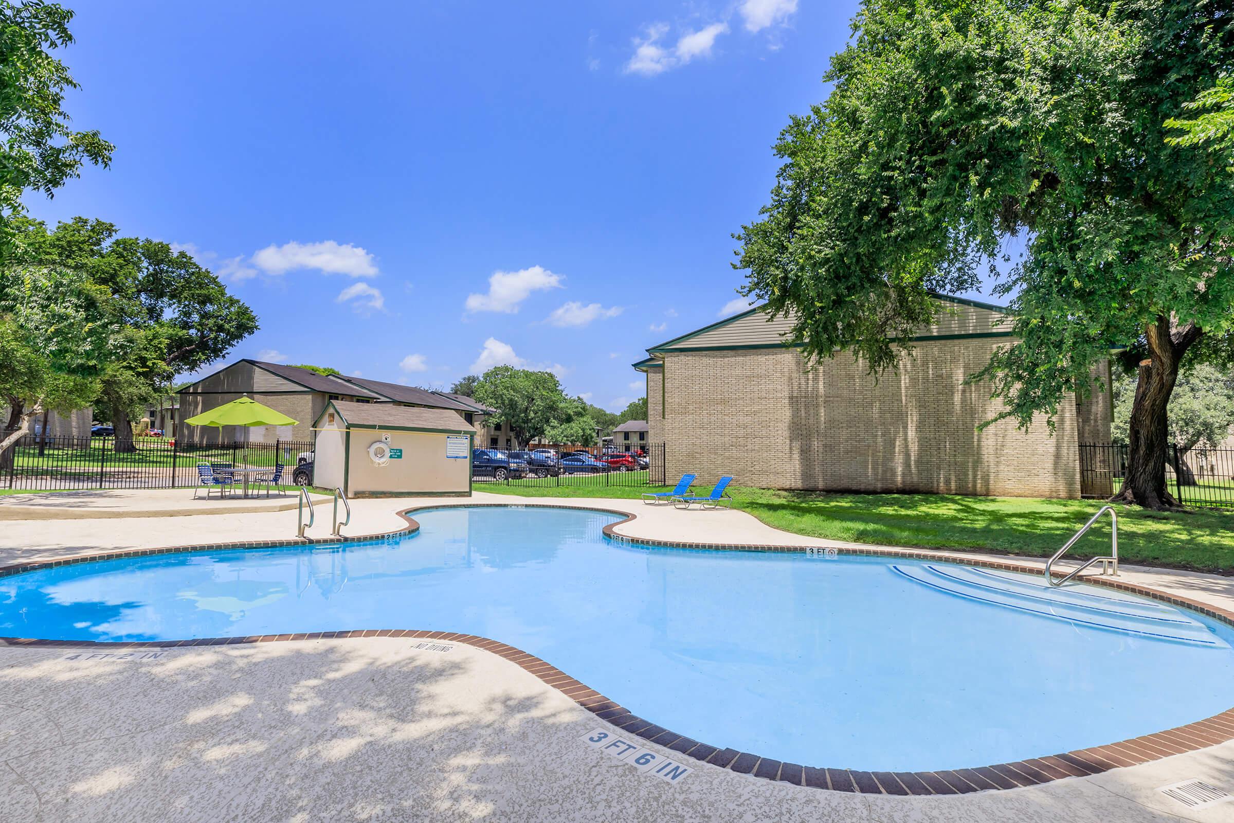 A clear blue swimming pool surrounded by greenery on a sunny day. Lounge chairs are positioned next to the pool. In the background, there are residential buildings and a shaded area with an umbrella. The scene conveys a relaxing outdoor atmosphere.
