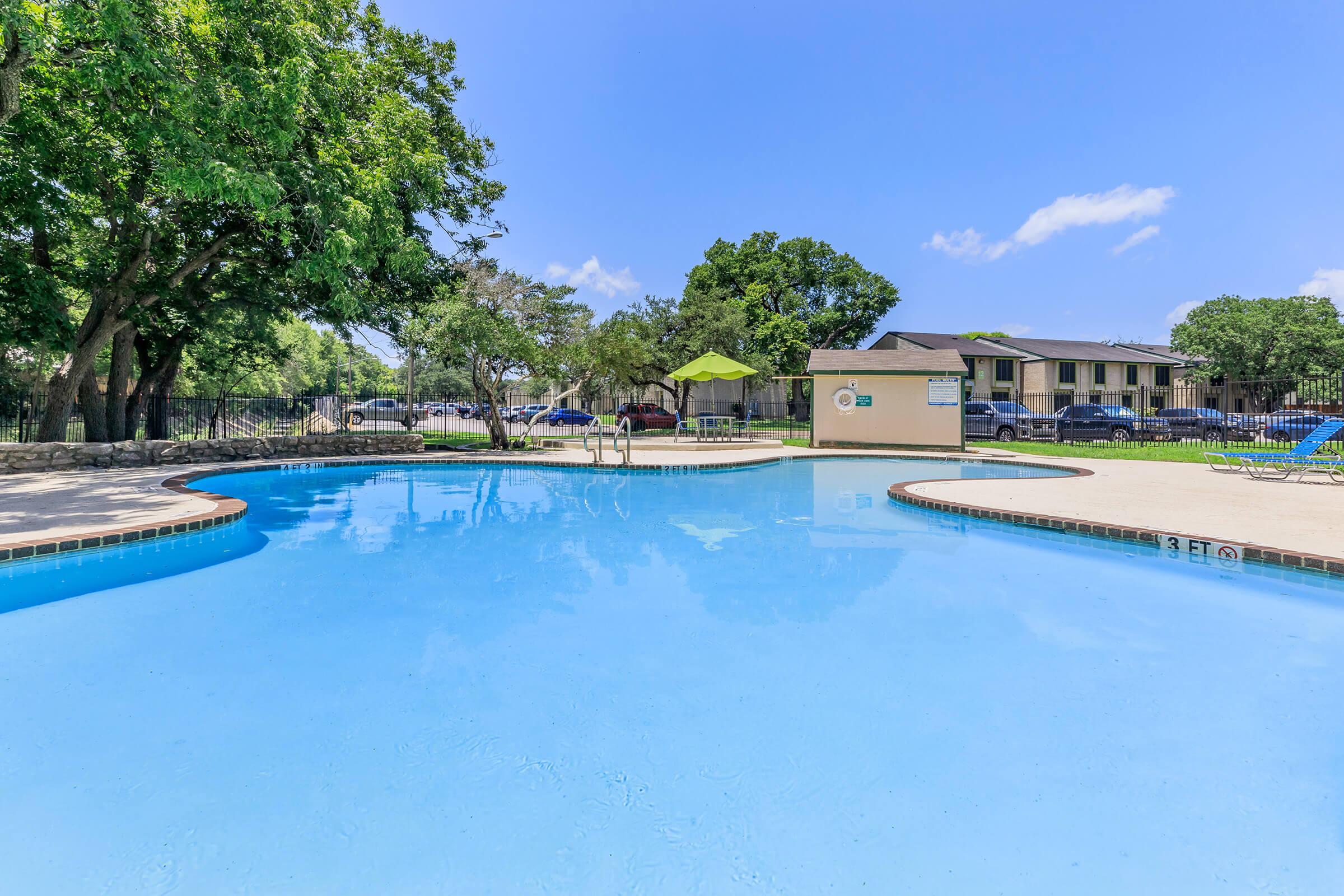 A sparkling blue swimming pool surrounded by lush greenery and trees. Sun loungers are arranged nearby, and a shaded area with an umbrella is visible. In the background, there are a few parked cars and an apartment building. The sky is clear and bright.